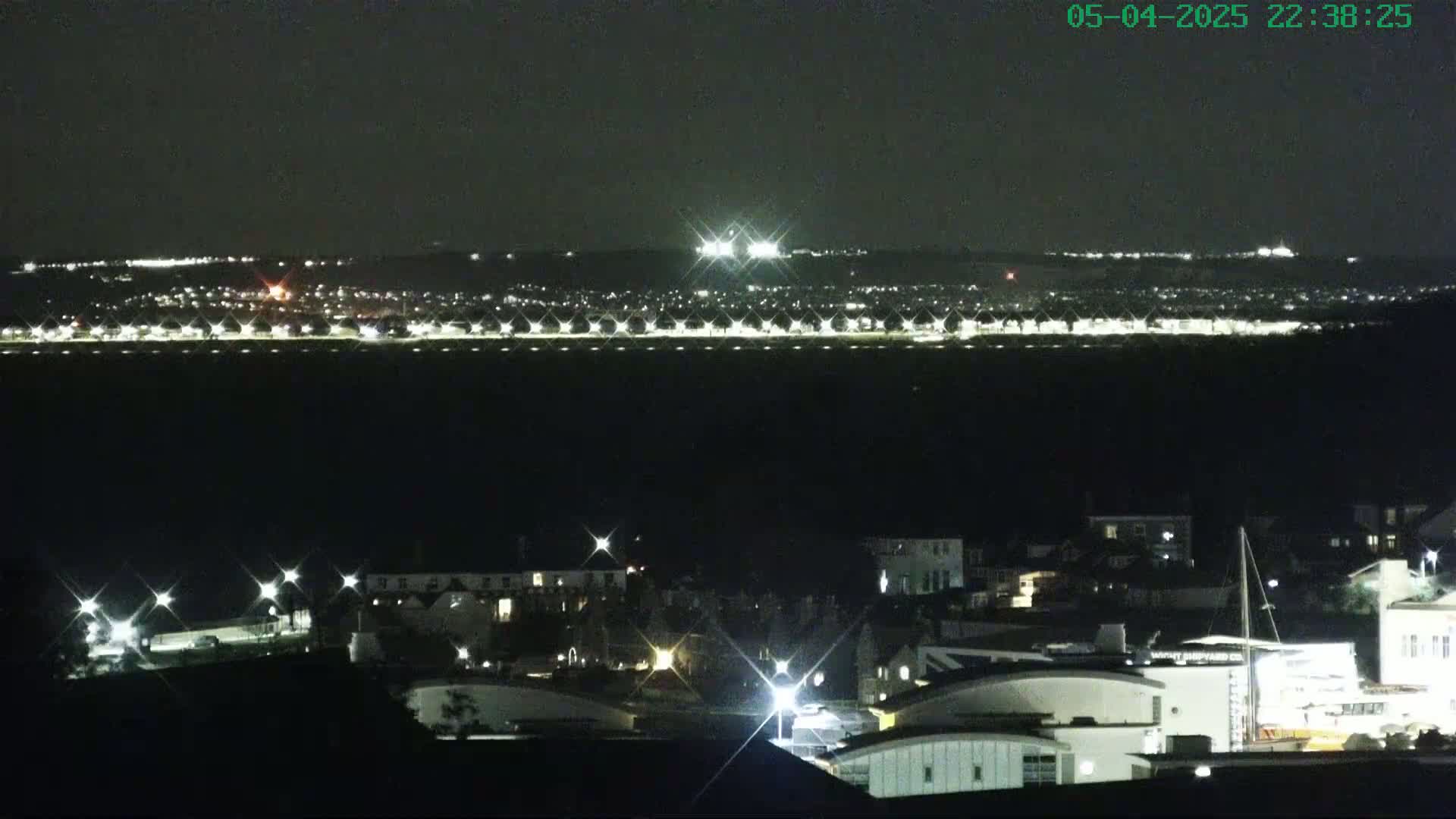 A nighttime view of a town's lights with a brightly lit area in the distance across a dark body of water.