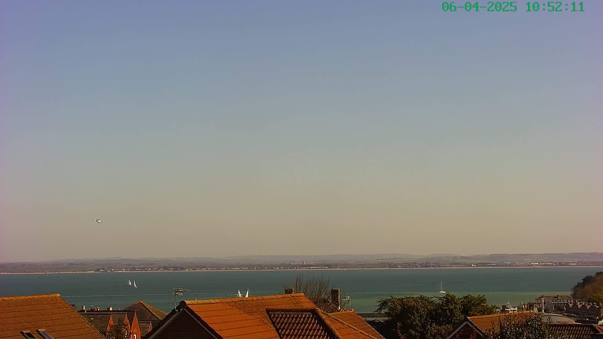 A calm, clear day reveals a wide expanse of teal water, dotted with a few sailboats, beyond a line of orange-tiled rooftops.