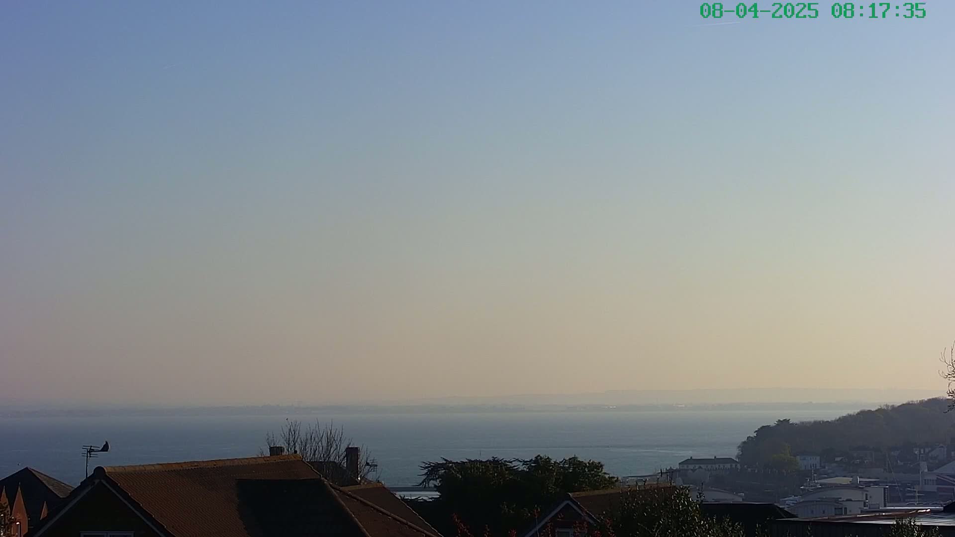 A hazy, clear day reveals a calm body of water extending to the horizon, beyond which lies a low-lying shoreline dotted with buildings and trees.