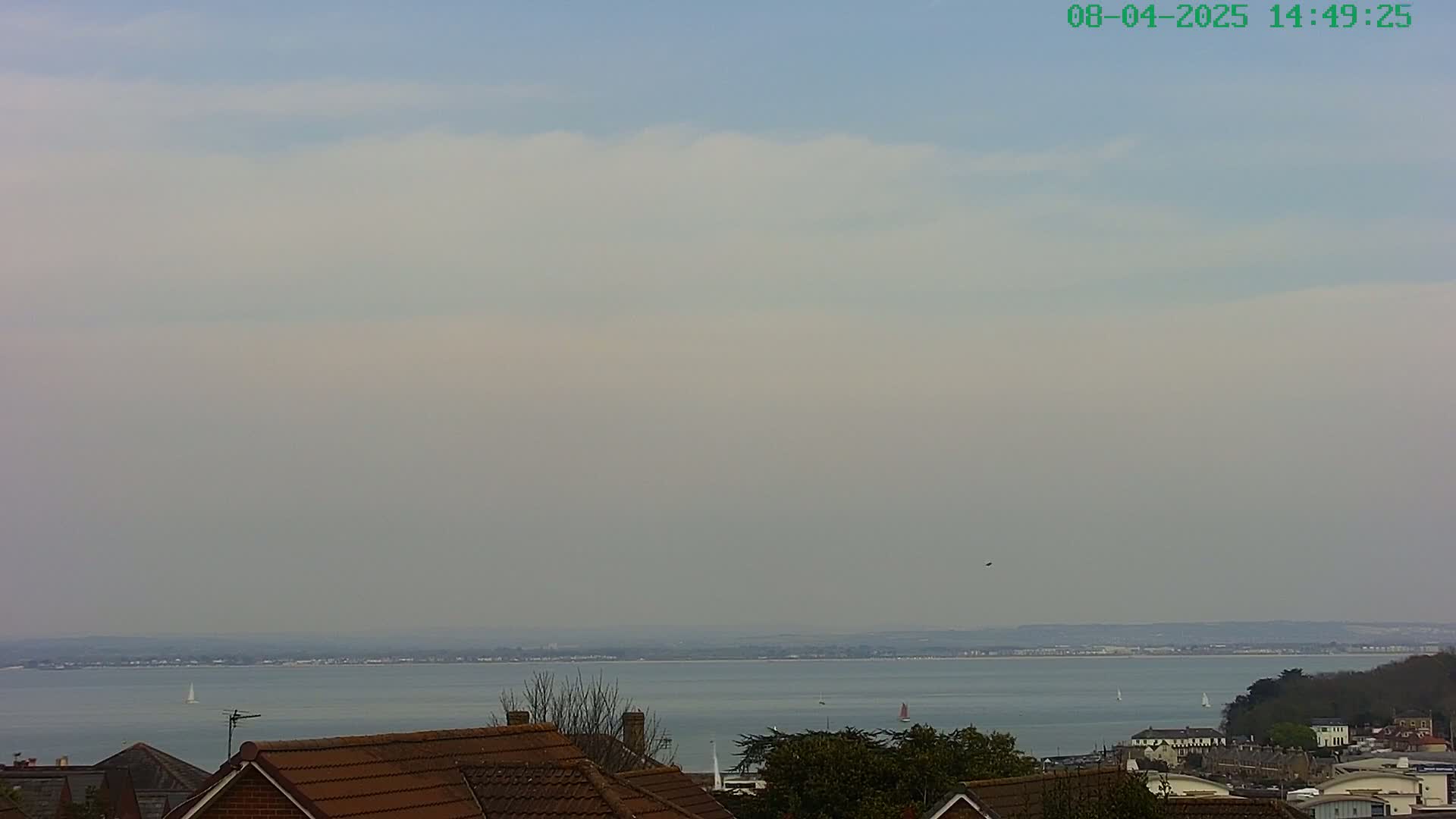 A hazy, mostly cloudy sky overlooks a calm body of water with a few sailboats, and rooftops of houses and trees are in the foreground.