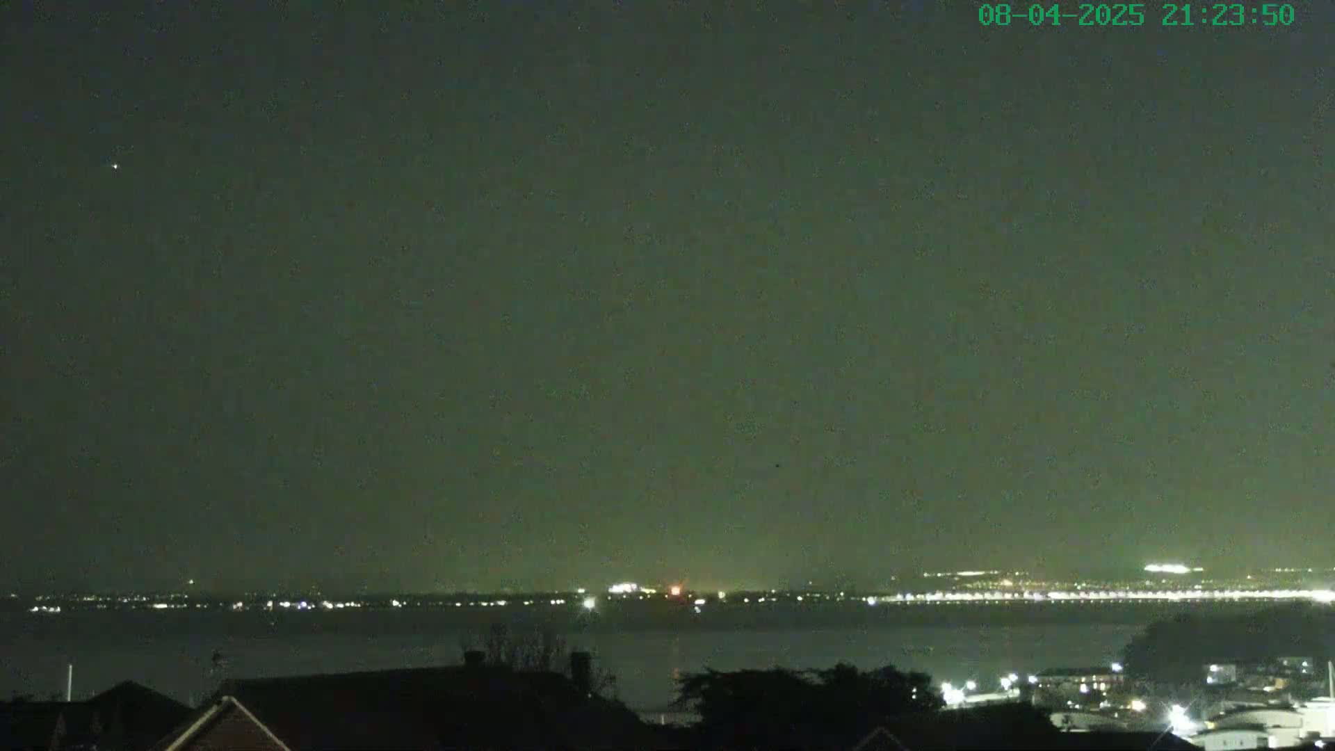 A nighttime view shows a body of water with a distant city skyline illuminated with lights under a dark, clear sky.