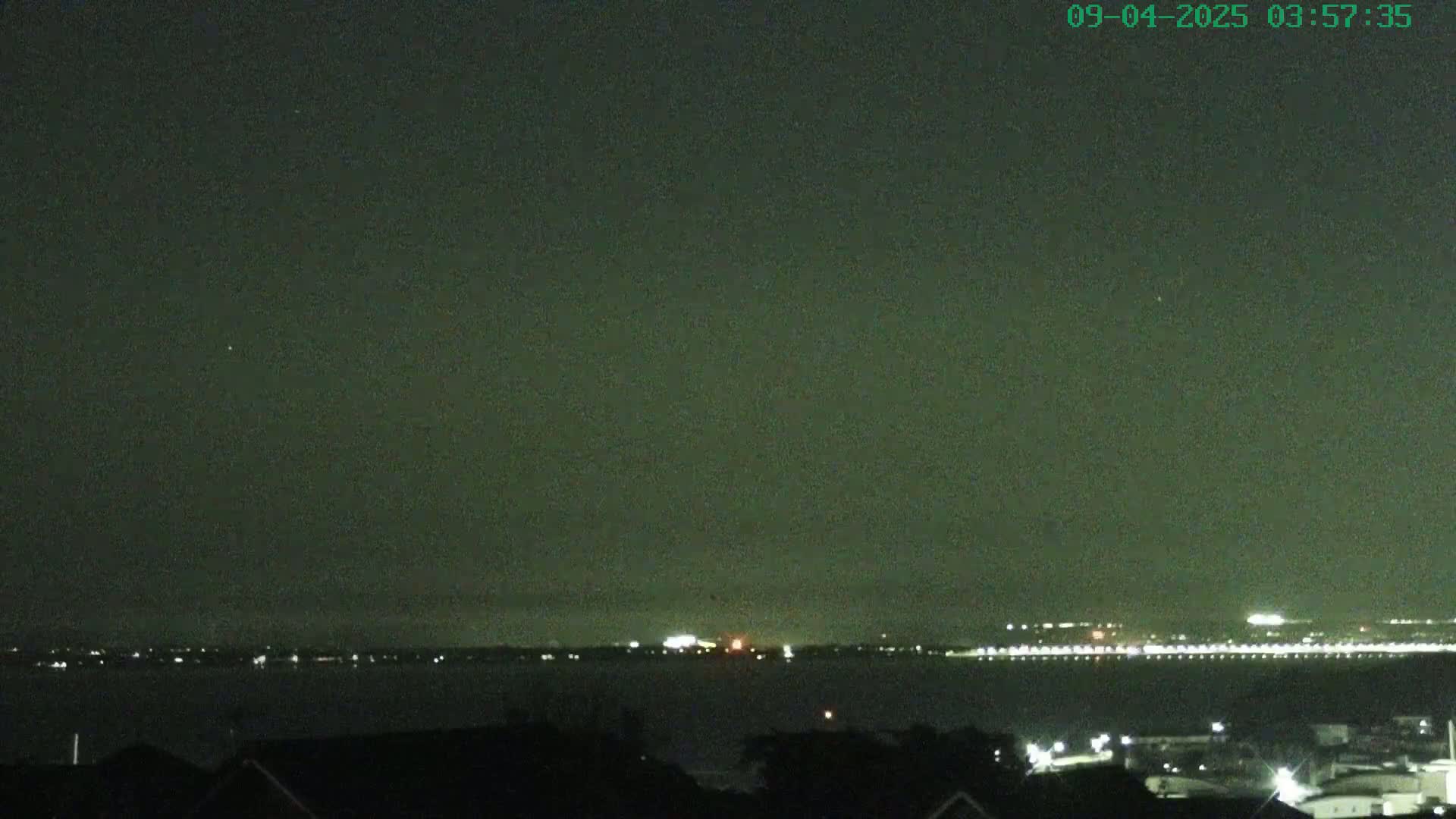 A nighttime view shows a dark body of water with a distant, faintly lit shoreline under a mostly clear, dark sky.