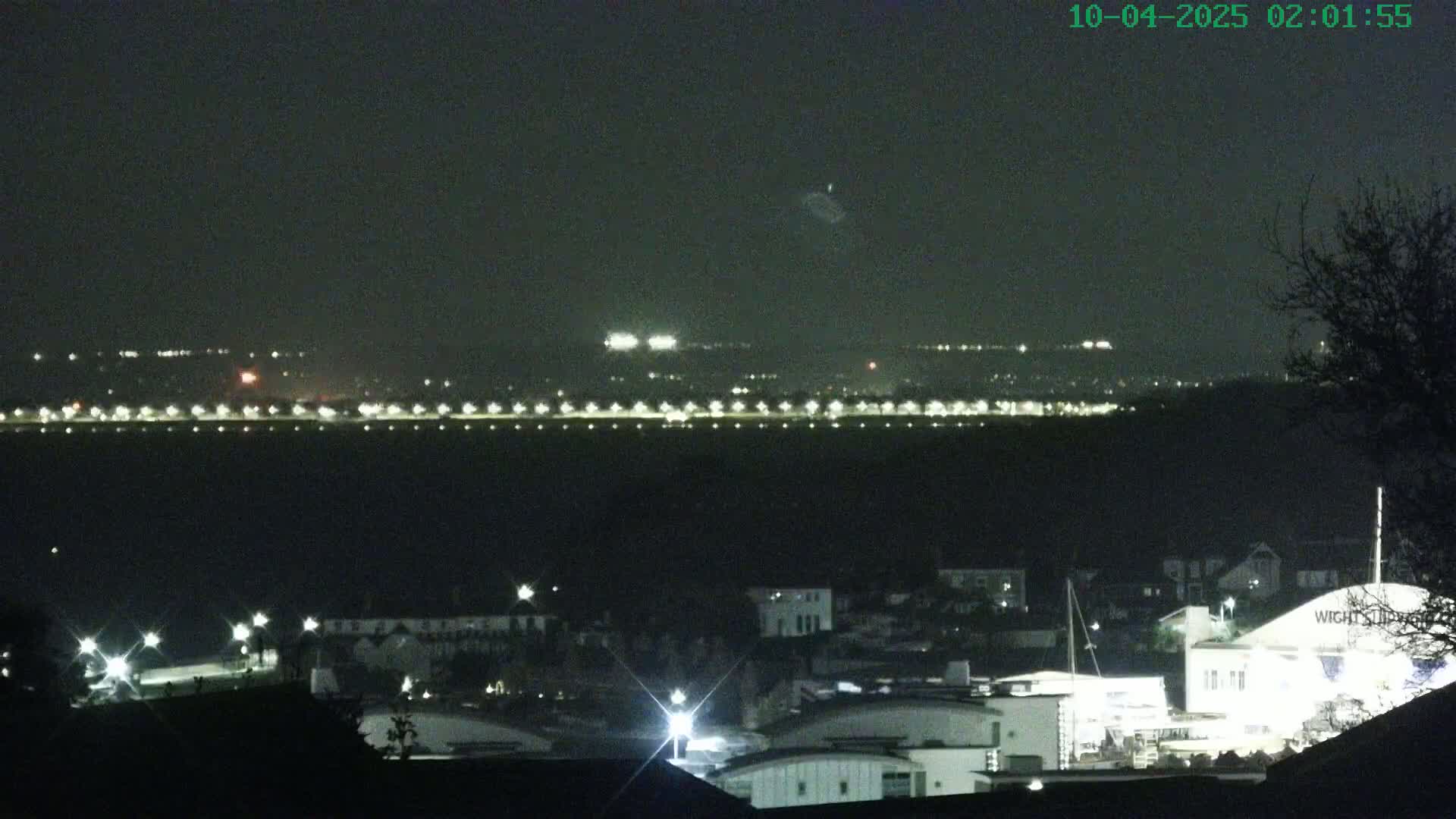 A nighttime long shot shows a town's illuminated waterfront and buildings, with a distant line of lights across the water under a dark, clear sky.