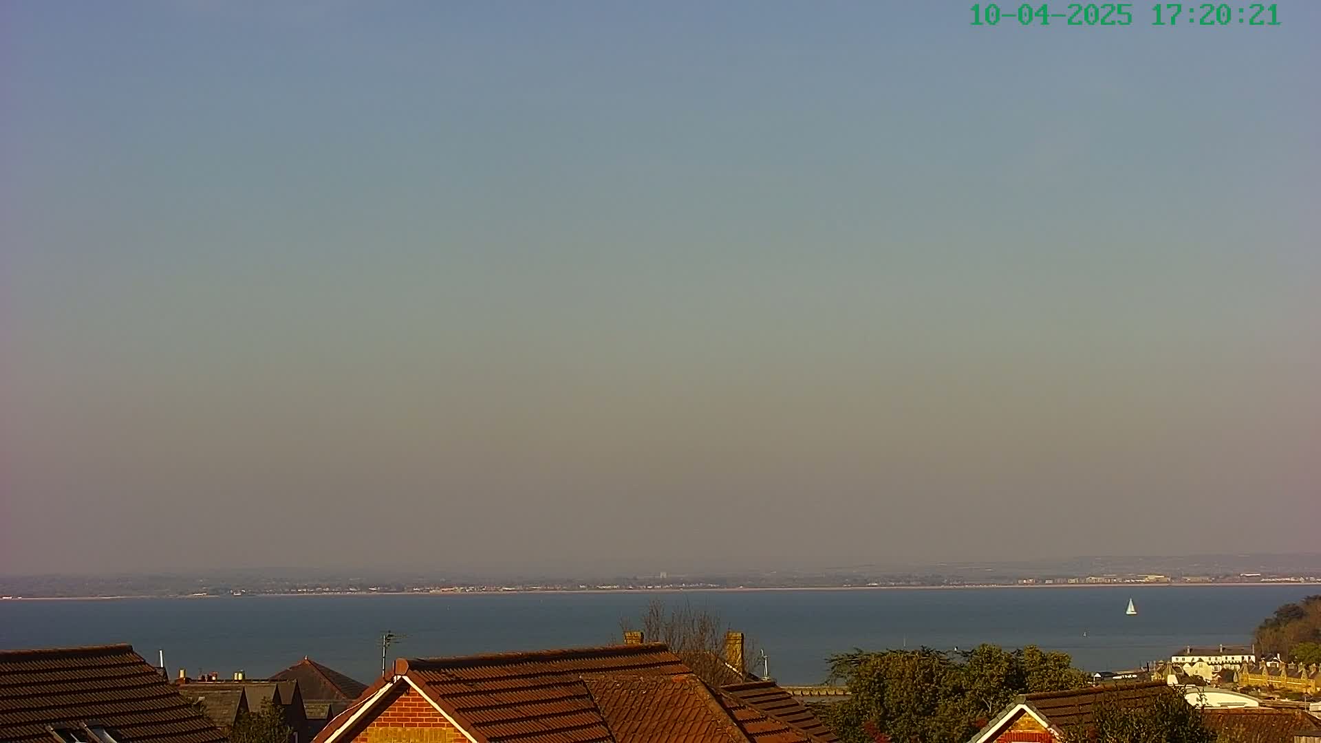 A hazy, calm day reveals a view of a calm body of water stretching to a distant shoreline, partially obscured by the rooftops of houses in the foreground.