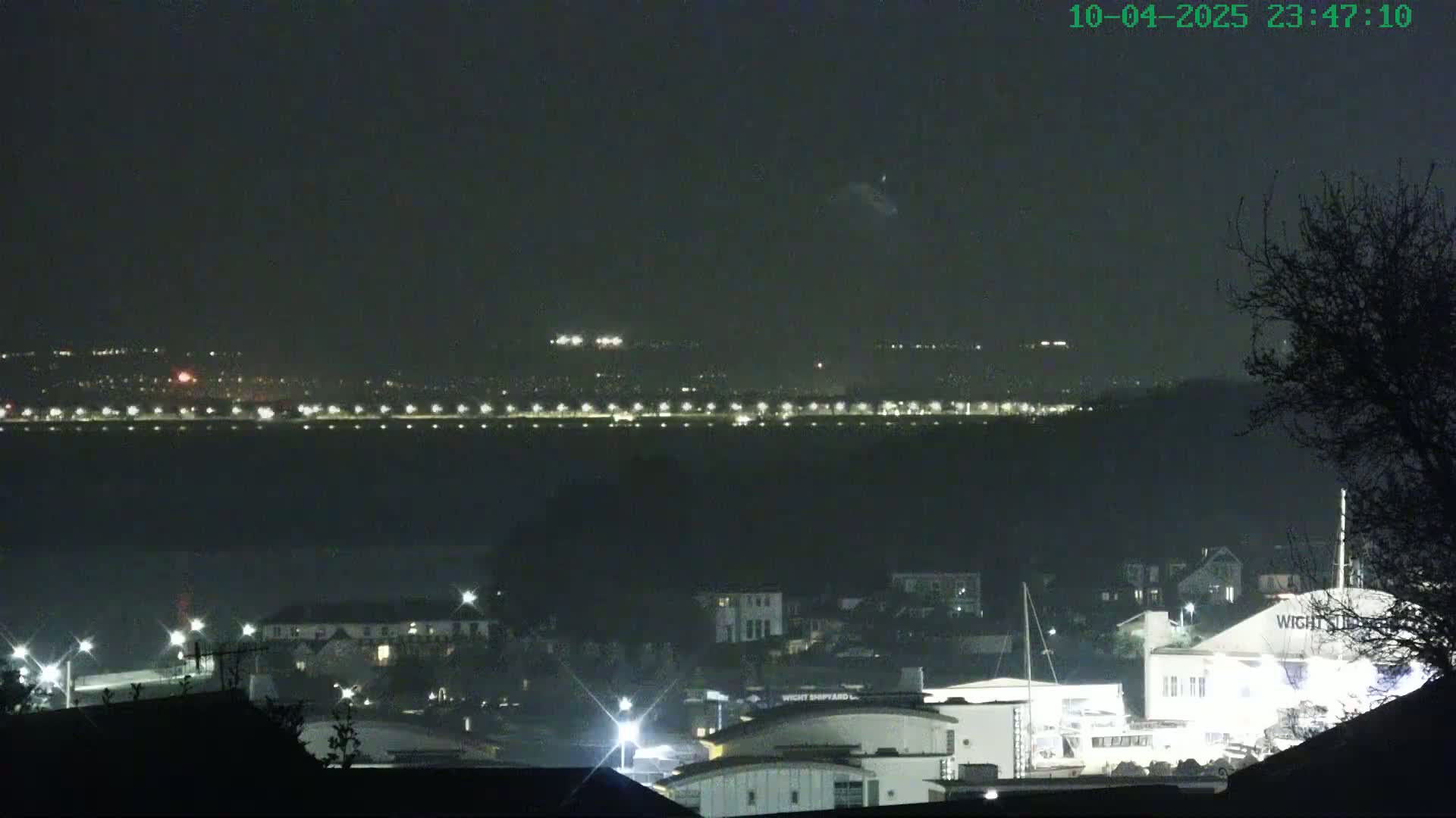 A nighttime view of a town illuminated by lights, with a distant, faintly lit coastline across a dark body of water under a clear, dark sky.