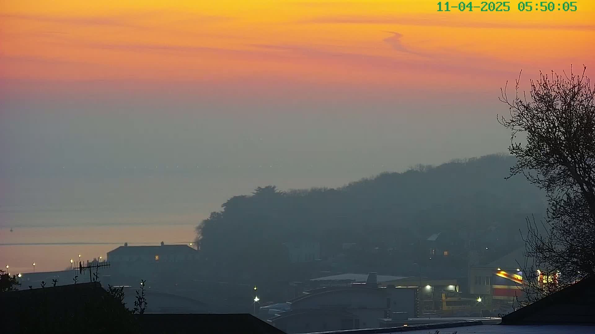 A hazy sunrise paints the sky in orange and pink hues over a distant body of water and a silhouetted town nestled among hills.