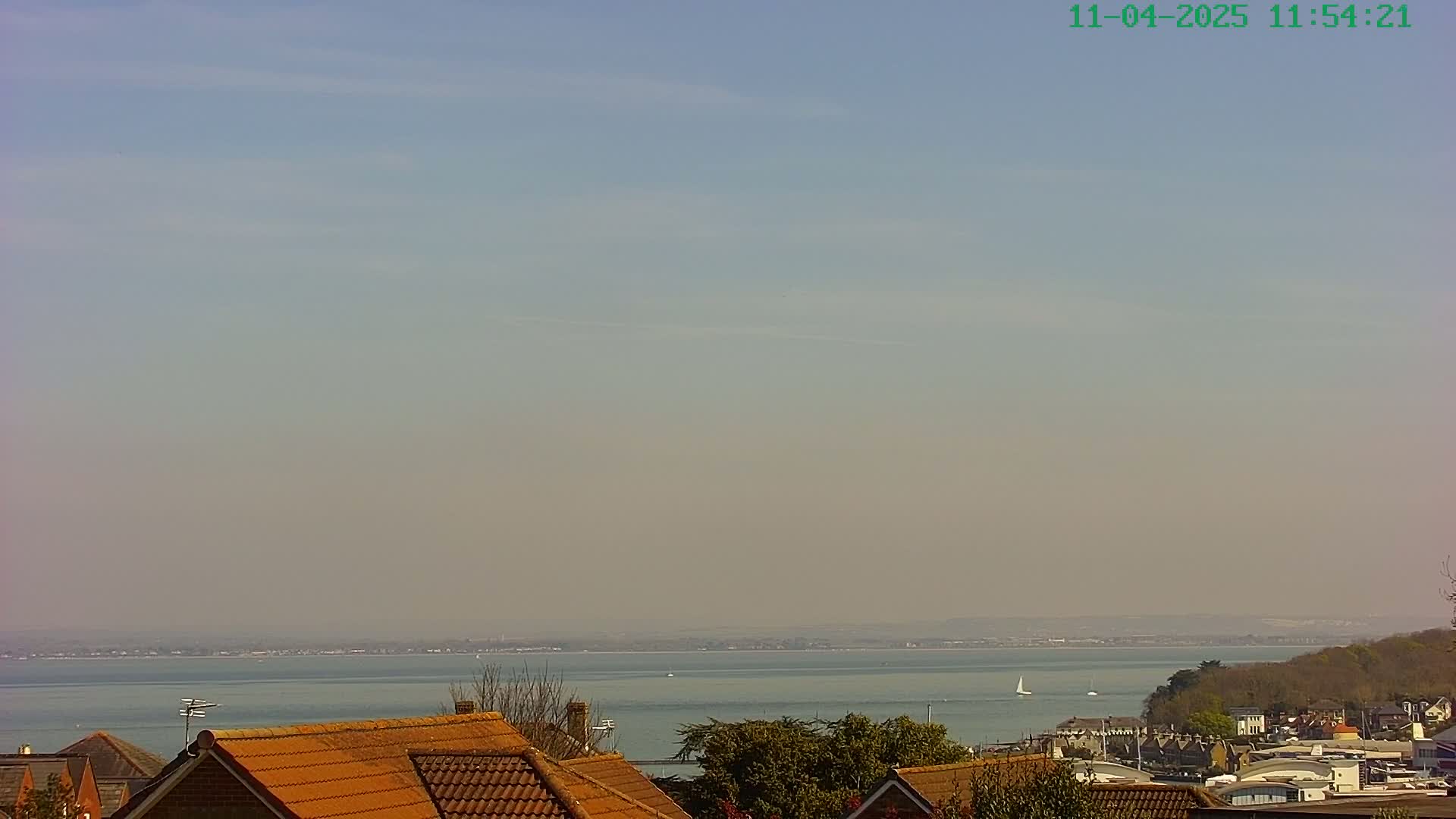 A hazy, sunny day overlooks a calm body of water with a distant shoreline and a few sailboats; in the foreground are the orange-tiled roofs of houses.