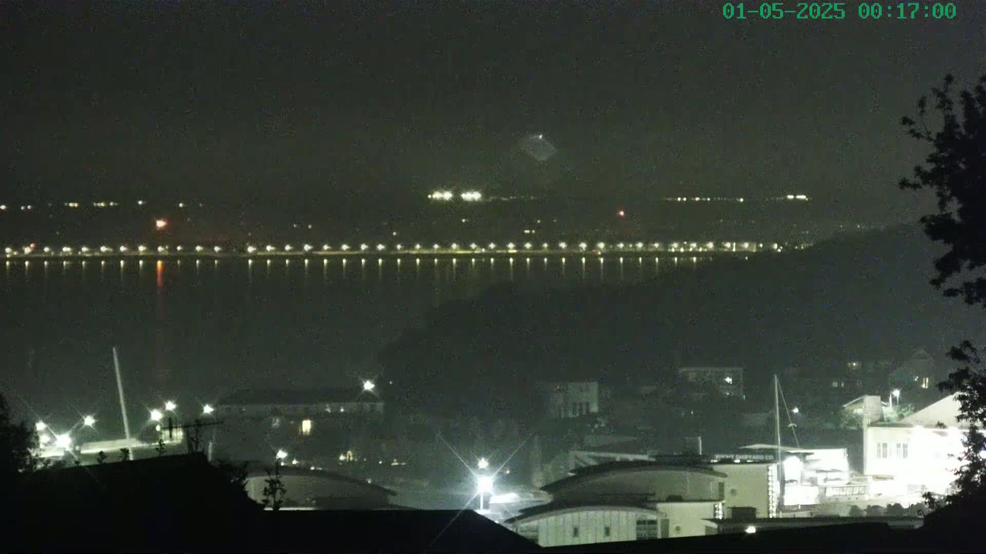 A nighttime view of a body of water with lights lining the far shore, and buildings and lights visible in the foreground under a clear, dark sky.