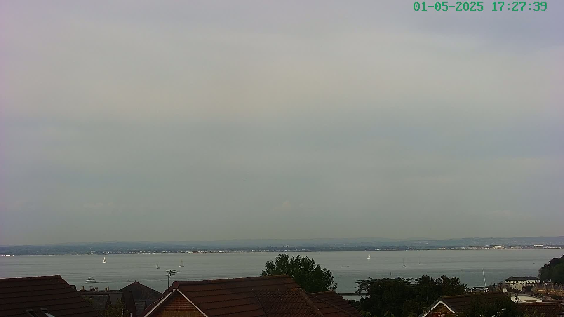 A hazy, overcast view shows a calm body of water with several sailboats, a distant shoreline, and the brown roofs of houses in the foreground.