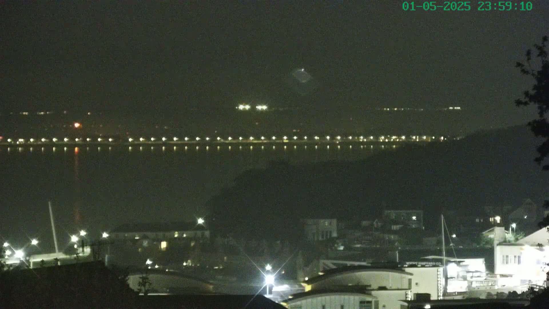 A nighttime view of a waterfront town with a body of water showing lights across a distant shore, under a hazy, dark sky.