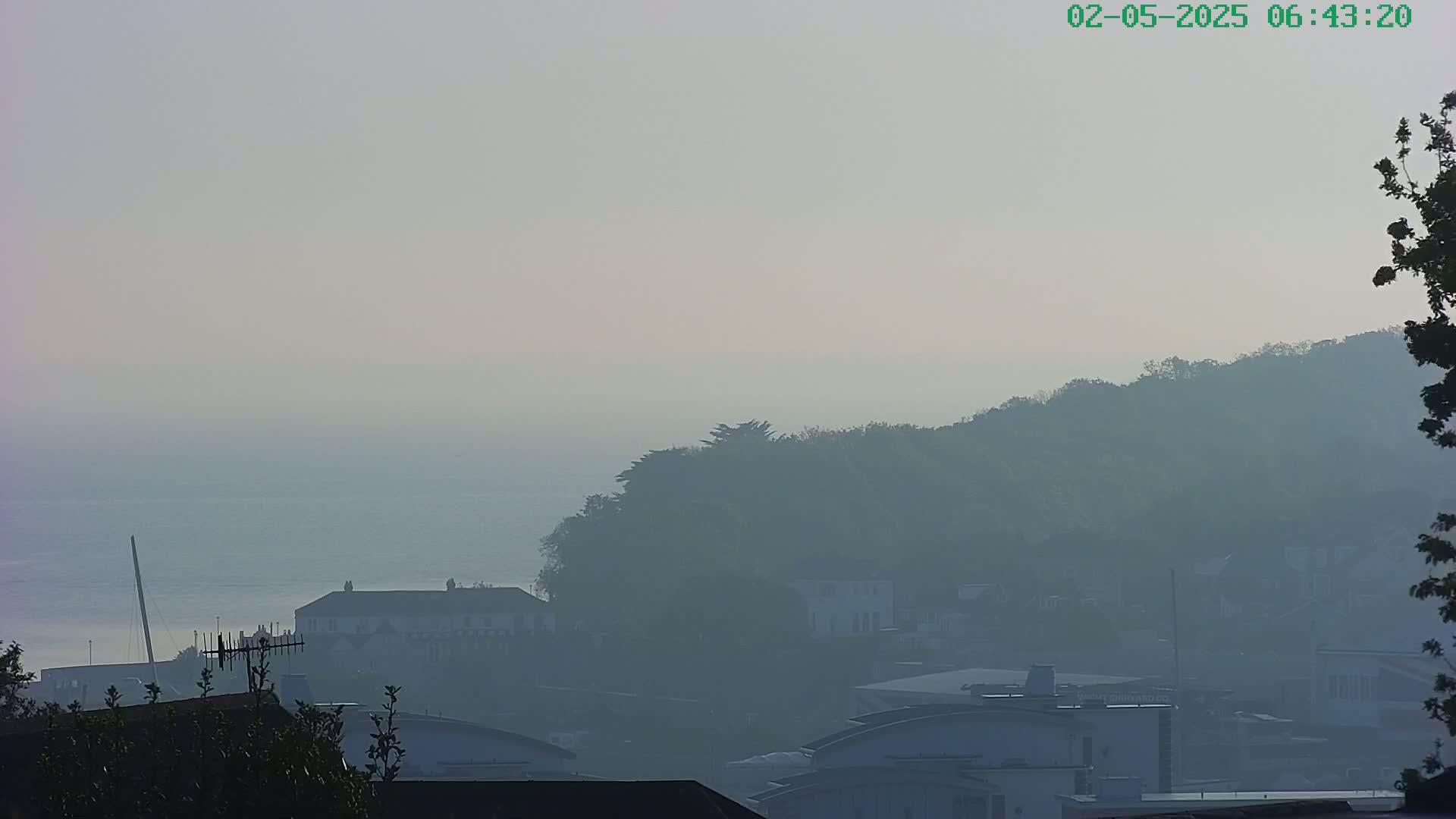 A hazy, early-morning view of a coastline shows buildings and trees obscured by fog or mist, with calm water visible in the distance.