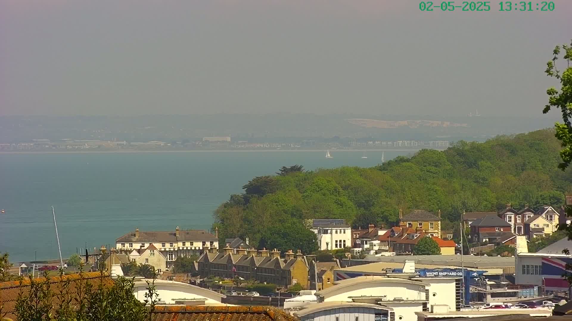 A hazy, sunny day overlooks a coastal town nestled beside a body of water, with green hills and distant land visible.