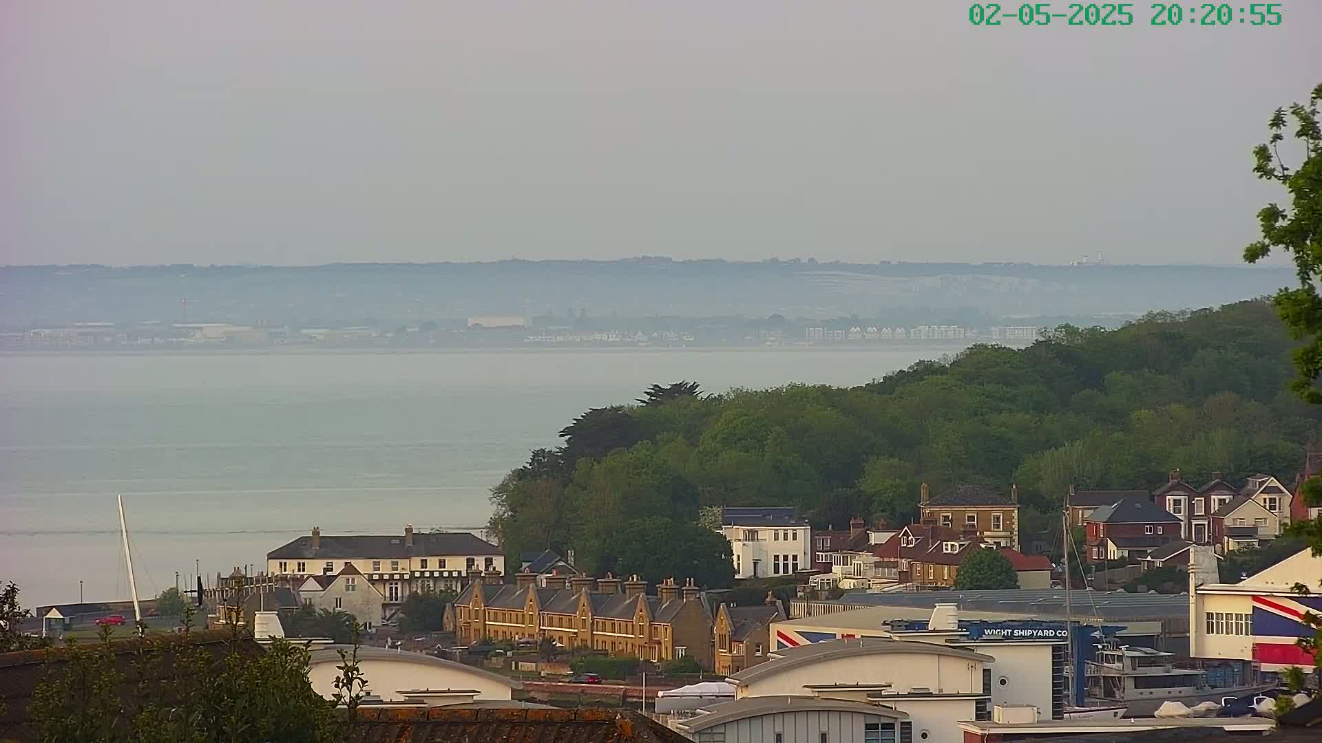 A hazy coastal town overlooks a calm body of water, with a line of buildings nestled amidst green trees.