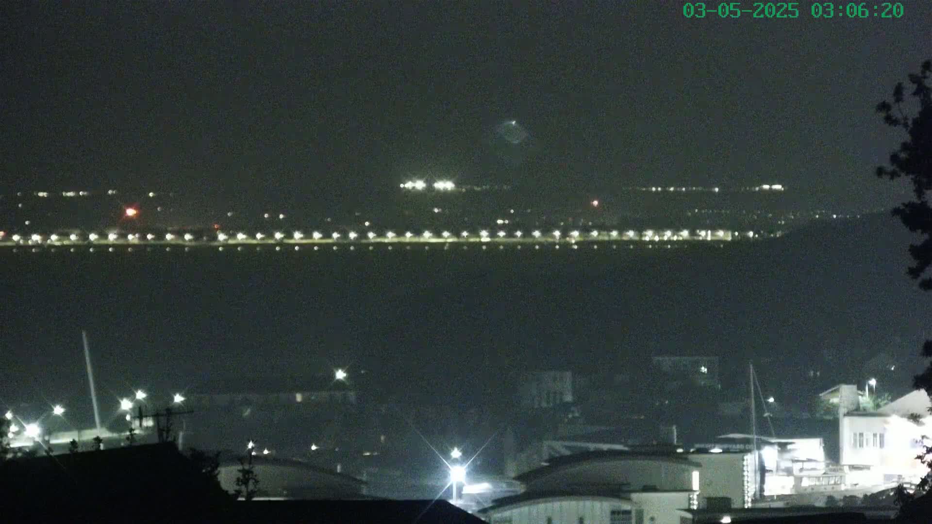 A nighttime view across a dark body of water shows a distant shoreline with lights, and a closer foreground with buildings and lights.