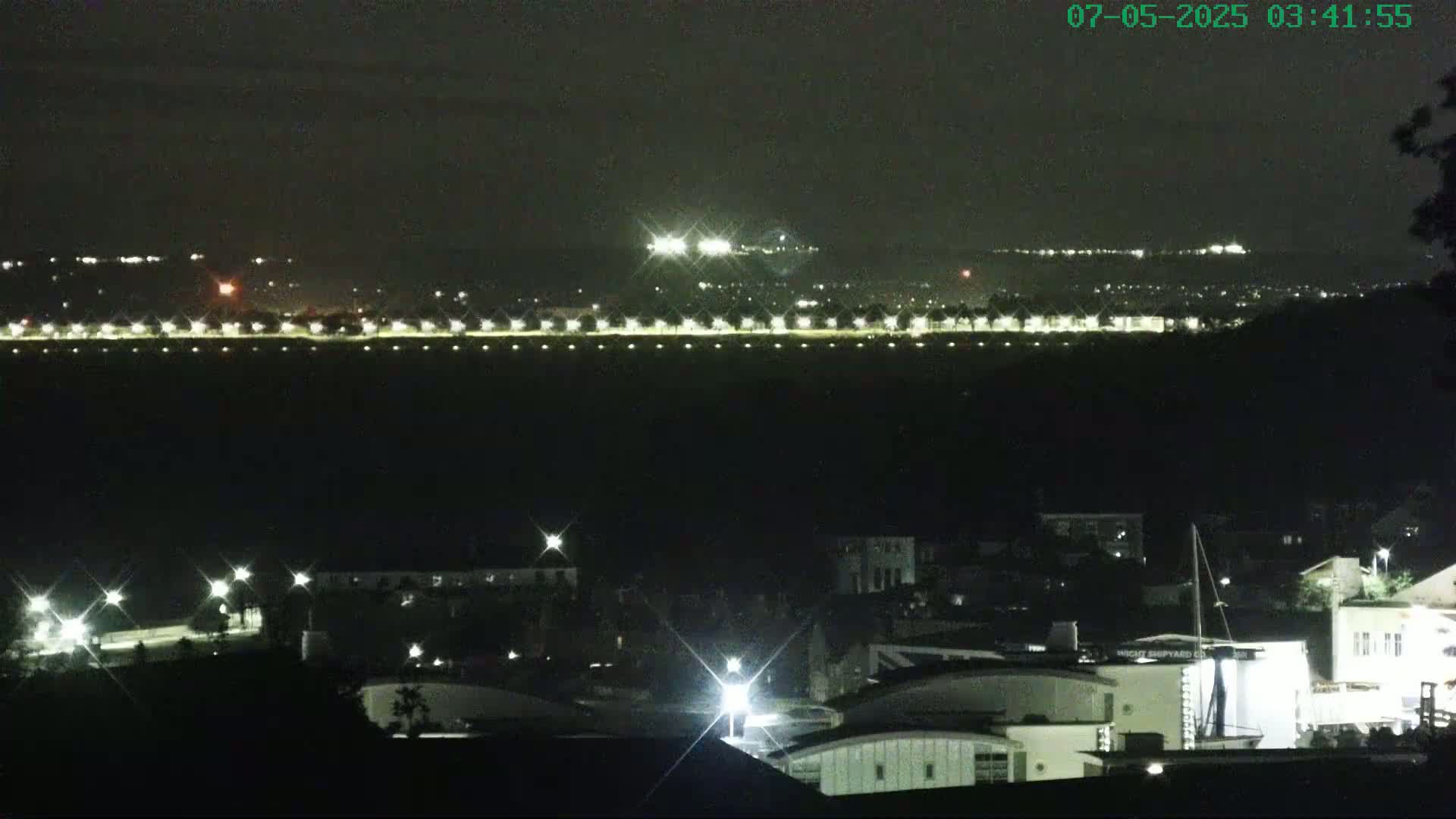 A nighttime view of a town with illuminated buildings and a distant, brightly lit area, possibly a highway or airport.
