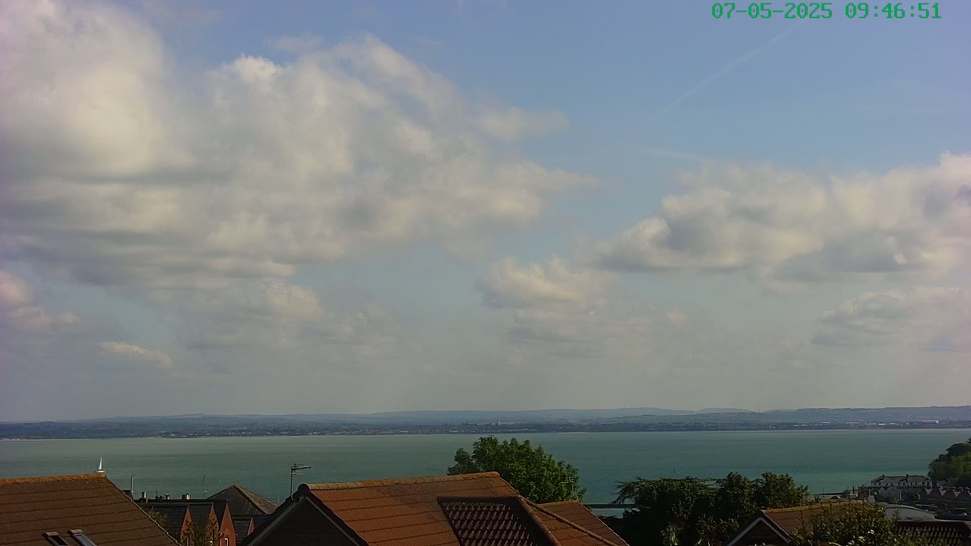A partly cloudy sky overlooks a calm body of water and a distant shoreline, with the tops of several brown-roofed houses visible in the foreground.