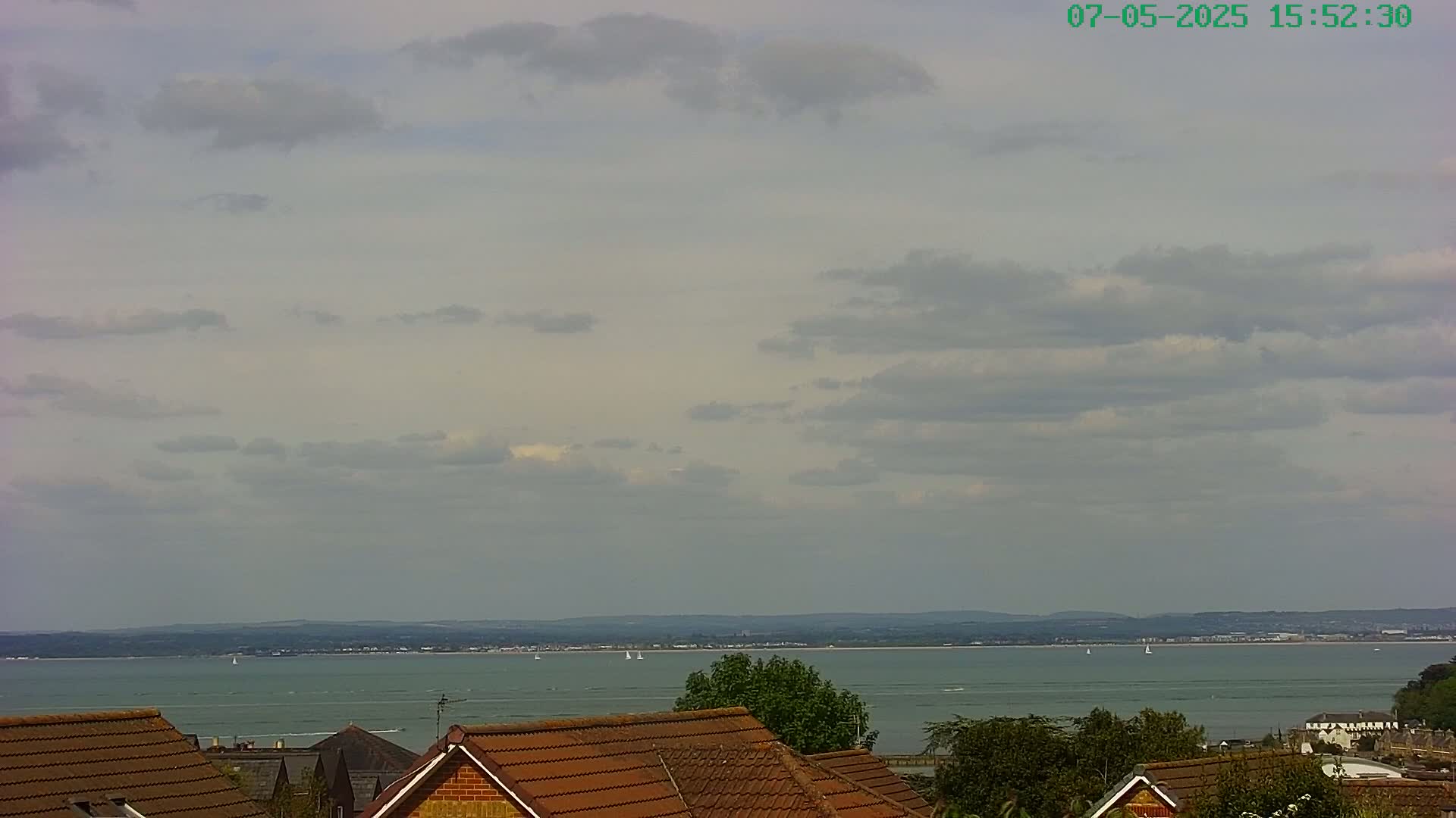 A partly cloudy sky overlooks a calm body of water with several sailboats, and a distant shoreline, viewed from behind the brown-tiled roofs of houses.