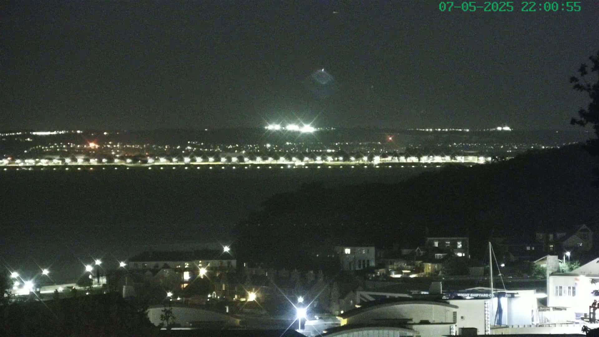 A nighttime view of a coastal town with a body of water separating it from a brightly lit shoreline in the distance under a dark sky.