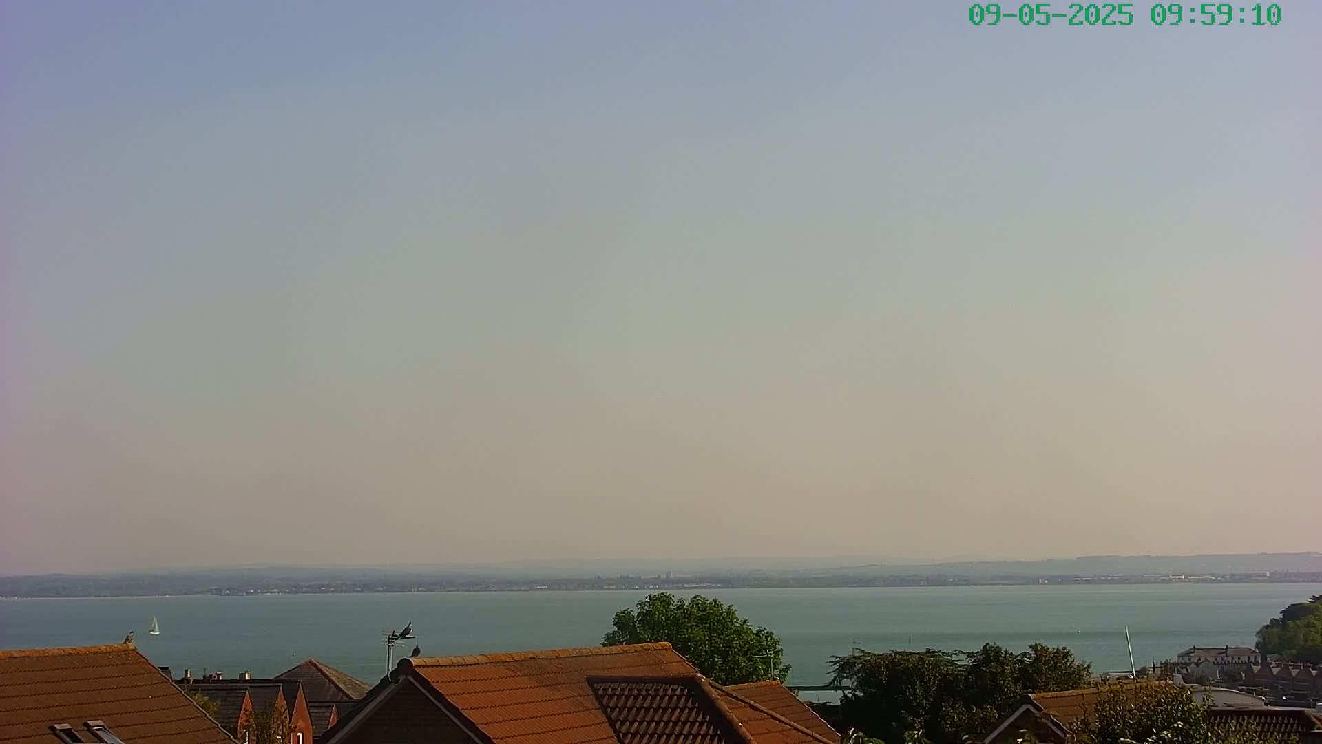 A hazy, sunny day overlooks a calm body of water from a vantage point above brown-tiled rooftops and green trees.
