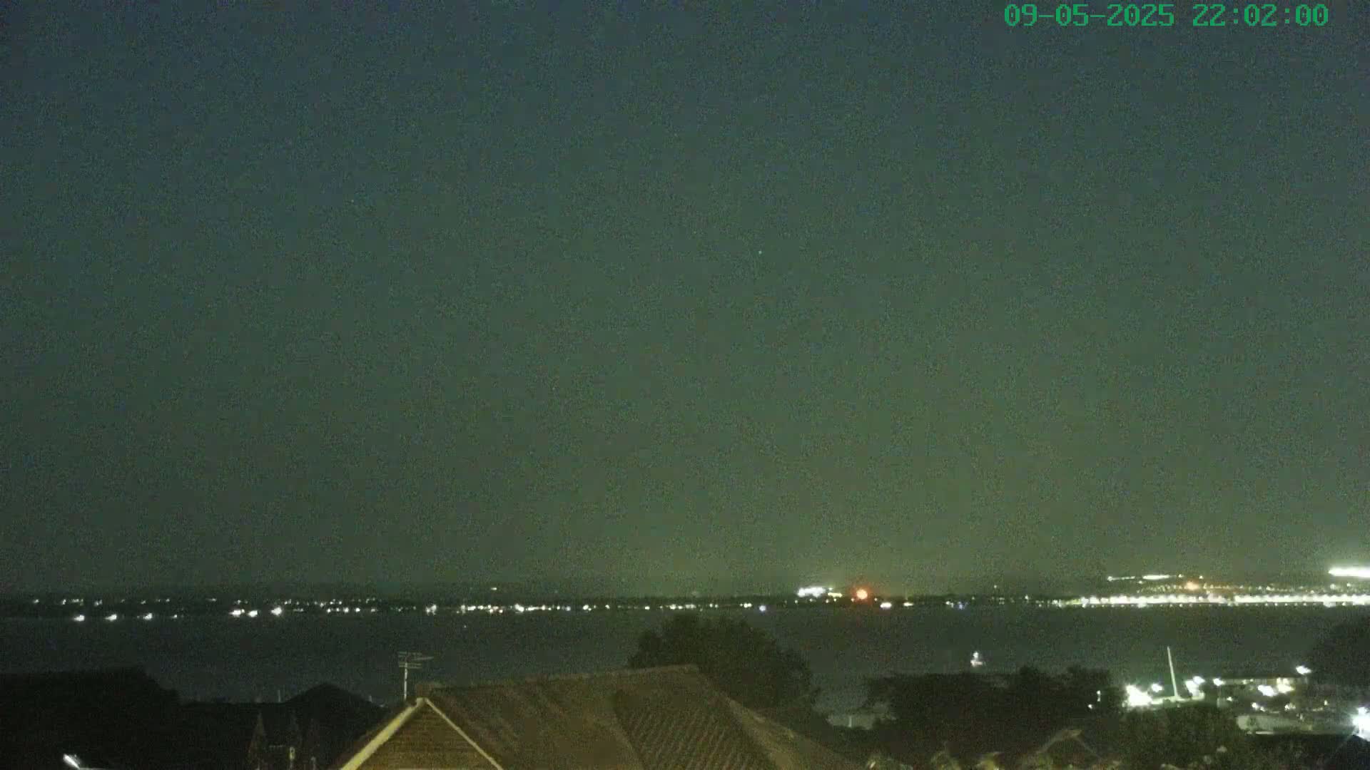 A nighttime view shows a dark body of water with a line of distant lights across the horizon, partially obscured by a hazy atmosphere, and a dark rooftop in the foreground.