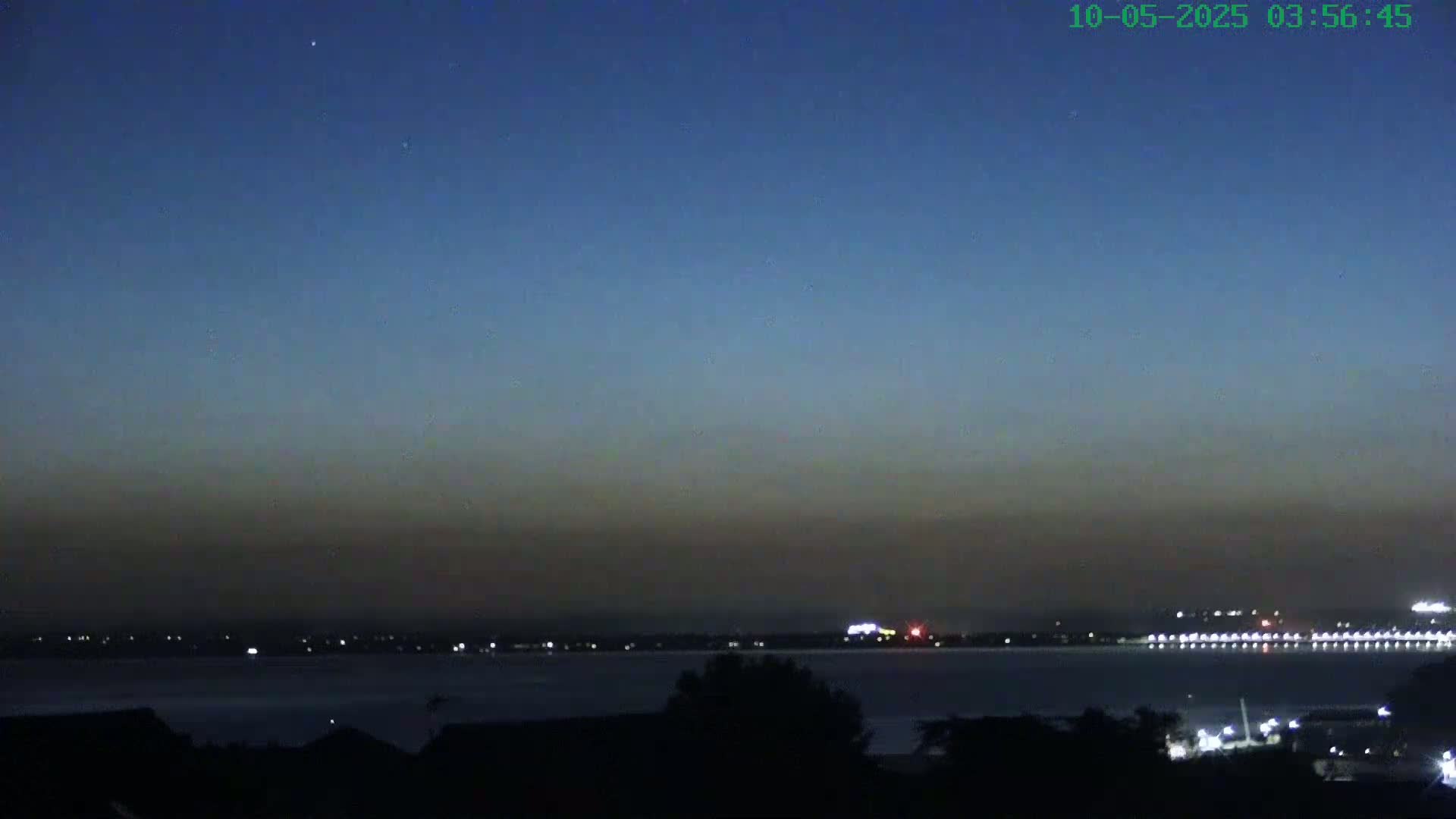 A dark, hazy twilight sky over a calm body of water with distant city lights and silhouetted trees in the foreground.