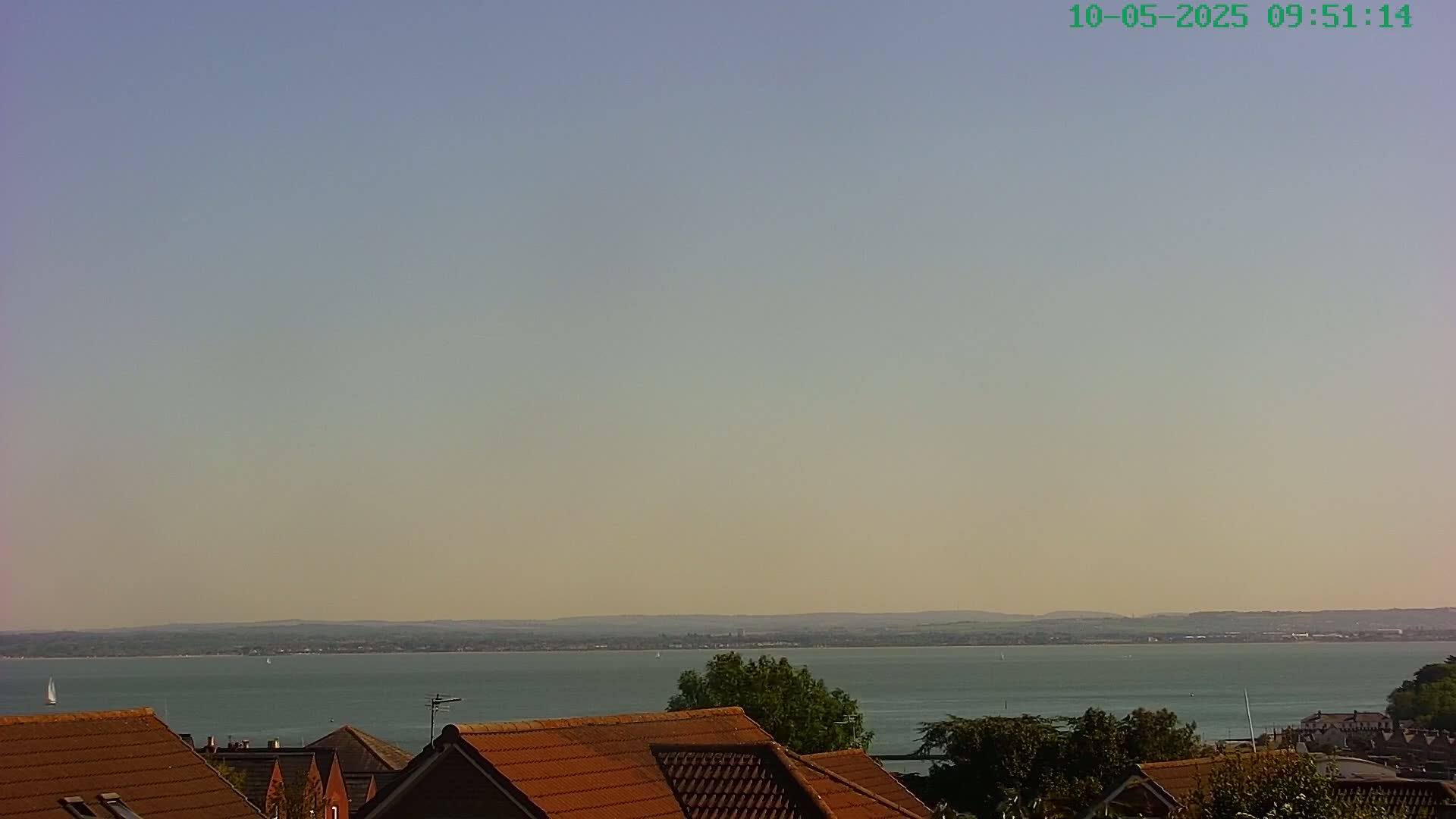 Under a clear, pale sky, a calm body of water stretches to a distant shoreline, viewed from above a row of brown-roofed houses.