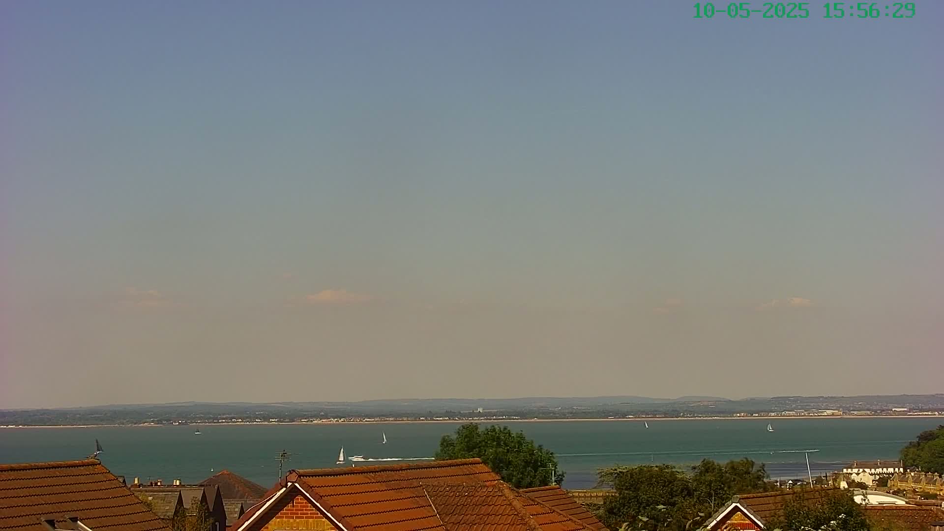 Under a mostly clear, light-blue sky, a calm body of water stretches to a distant shoreline, viewed from behind the reddish-brown roofs of several houses partially obscured by trees.