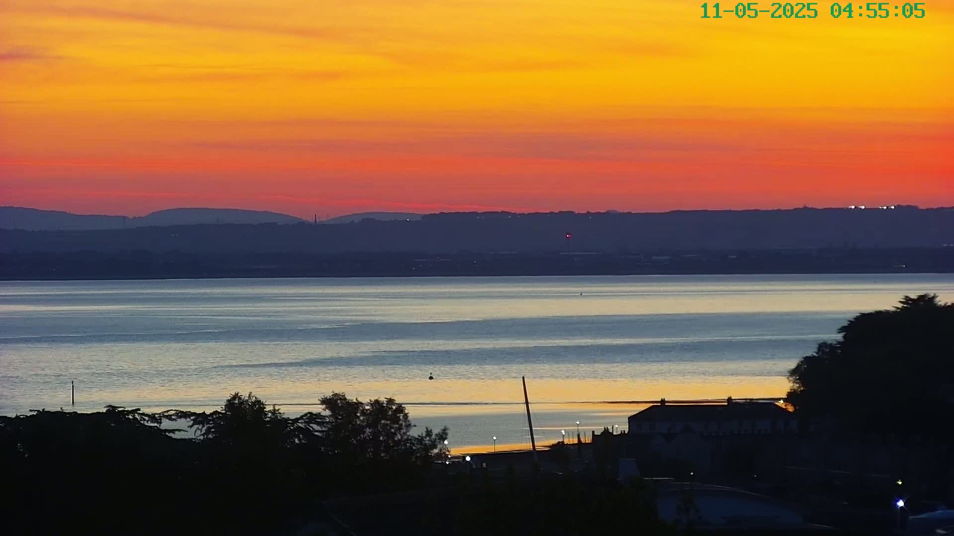 A fiery sunset over a calm body of water, silhouetting distant hills and a shoreline with buildings and trees.