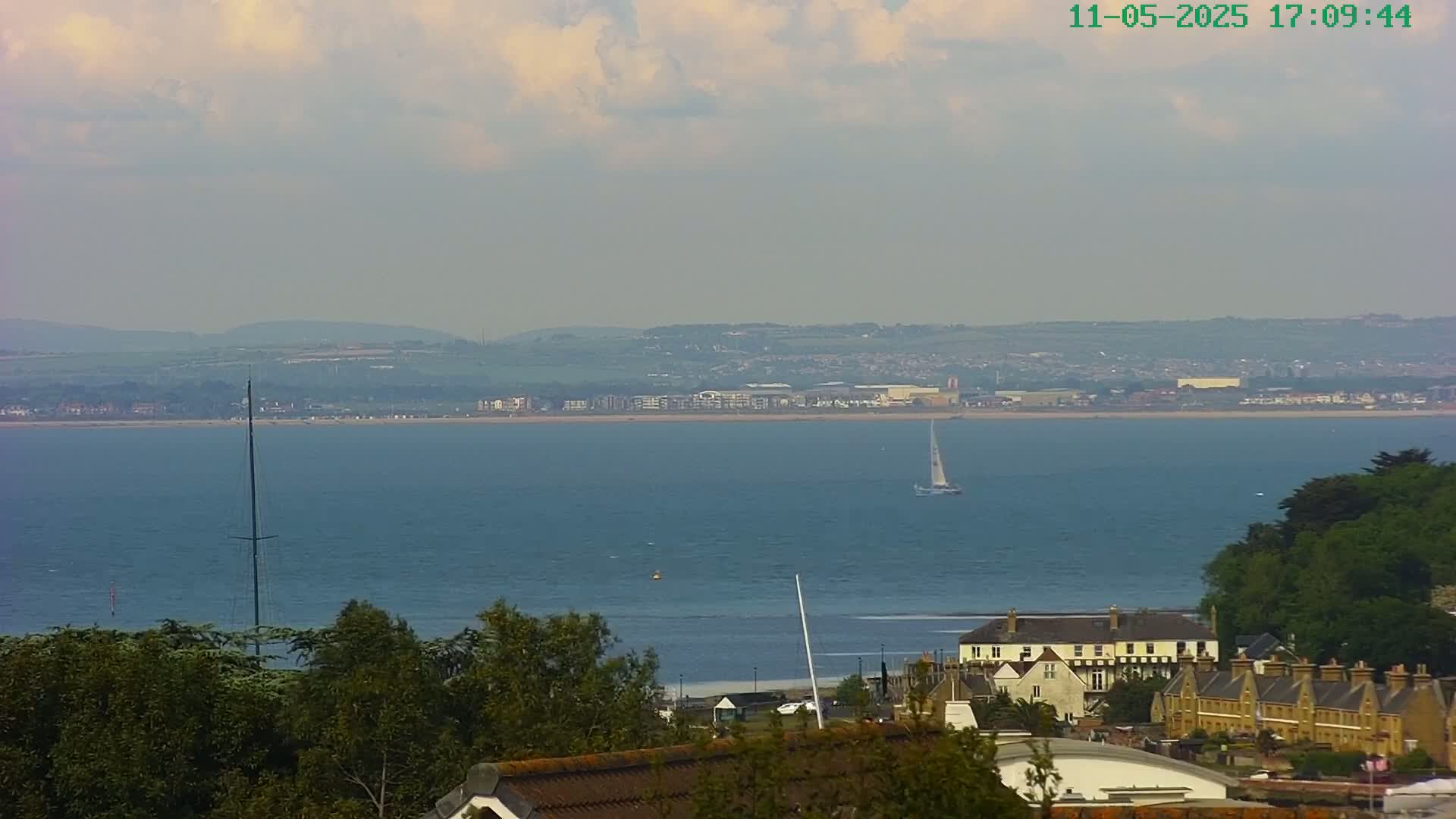 A sailboat is visible on a calm, blue body of water, with a distant shoreline featuring buildings and green hills under a partly cloudy sky.