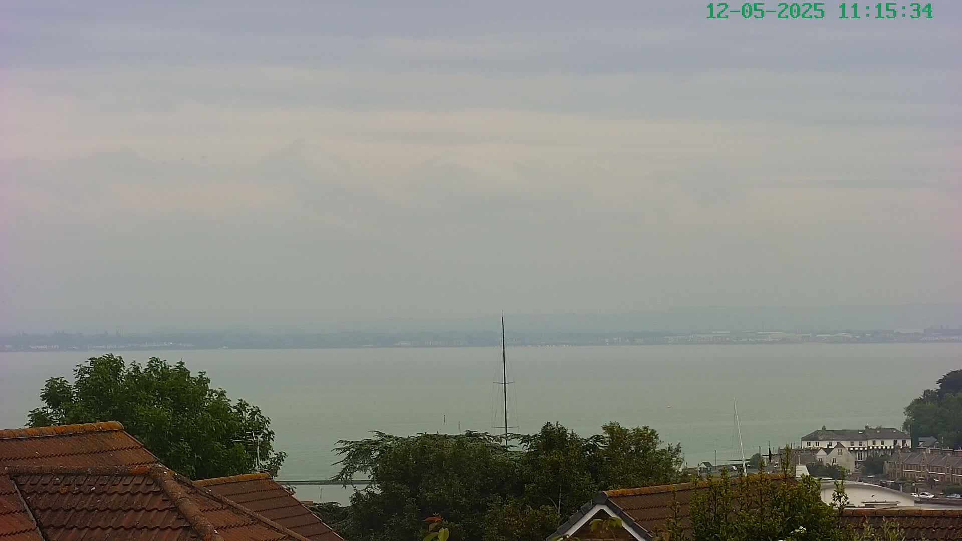 A hazy, overcast view shows a body of water with a sailboat in the distance, beyond houses with red-tiled roofs and green trees.