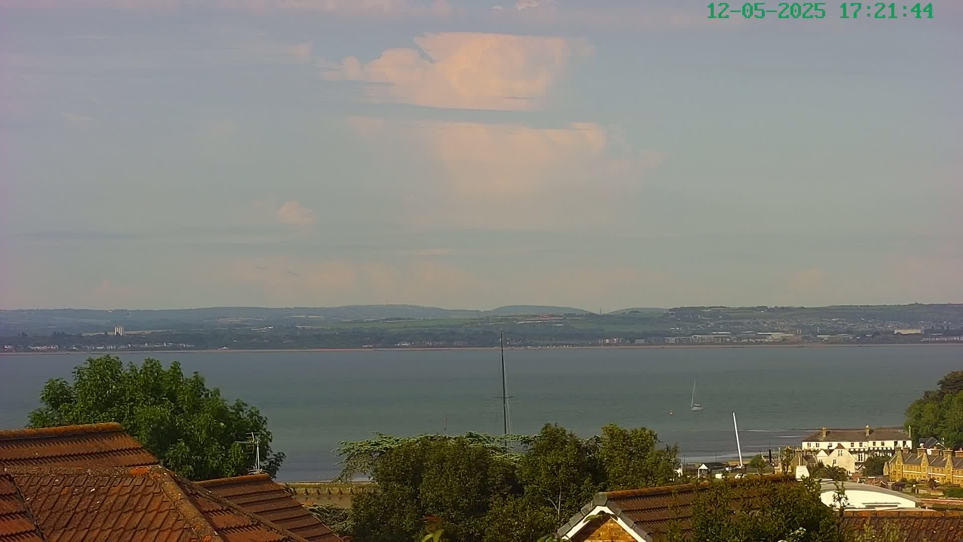 A partly cloudy sky overlooks a calm body of water, with distant land visible across the water and rooftops and trees in the foreground.