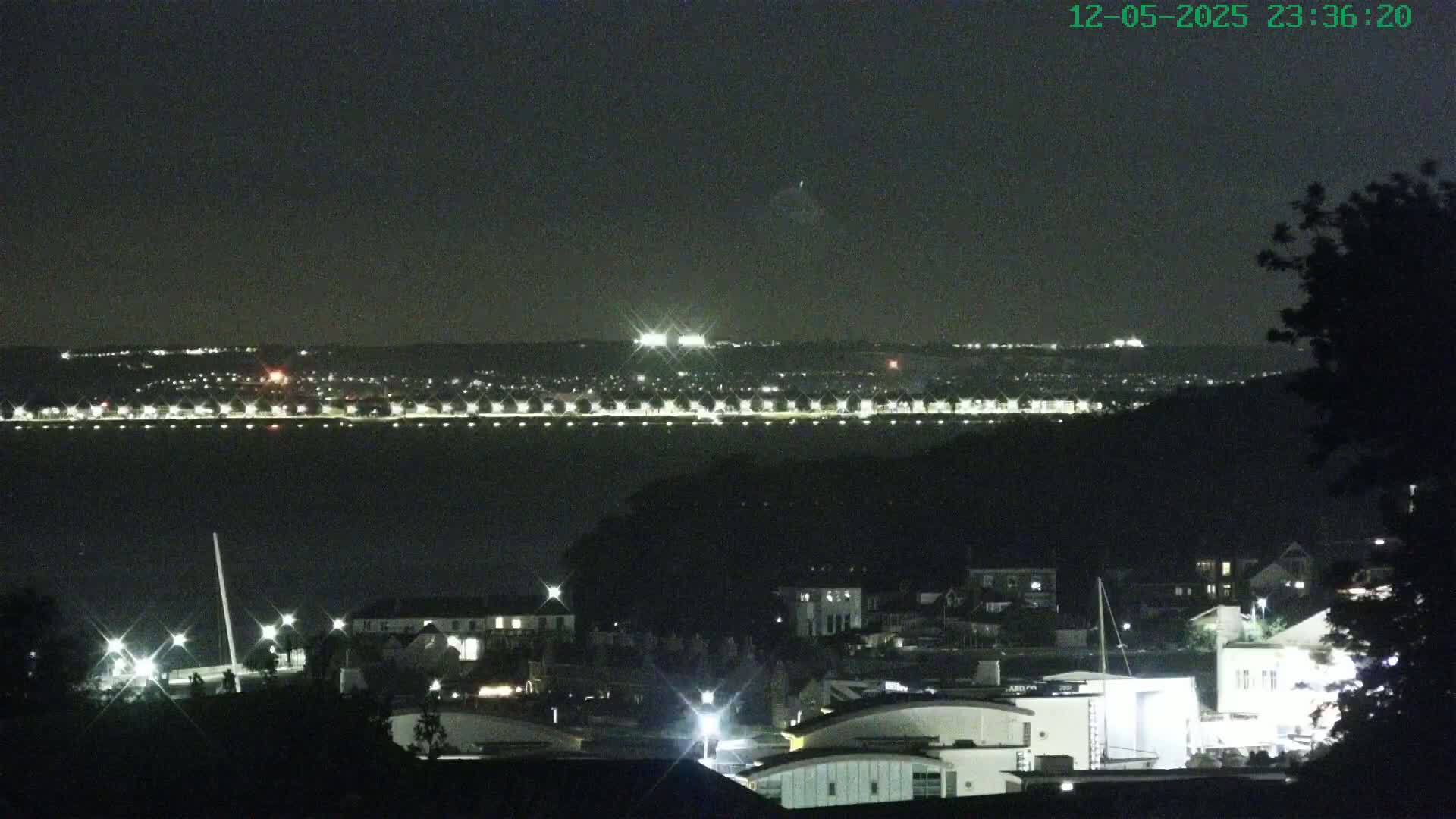 A nighttime view of a waterfront town illuminated by lights, with a darker, hilly area in the foreground and a distant, brightly lit shoreline across a body of water.