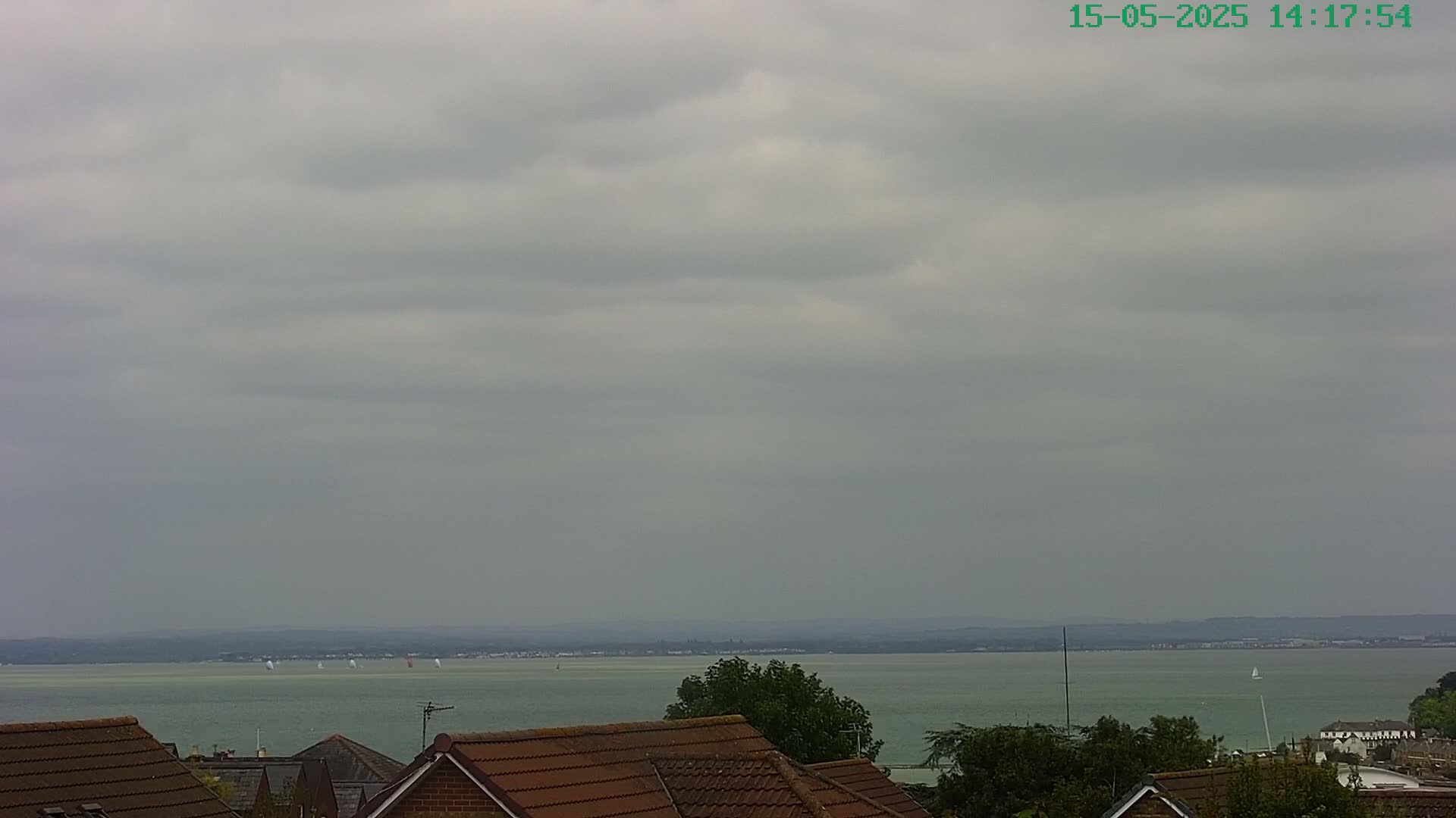 A view of a calm body of water under an overcast sky, with several sailboats visible in the distance and the tops of houses in the foreground.