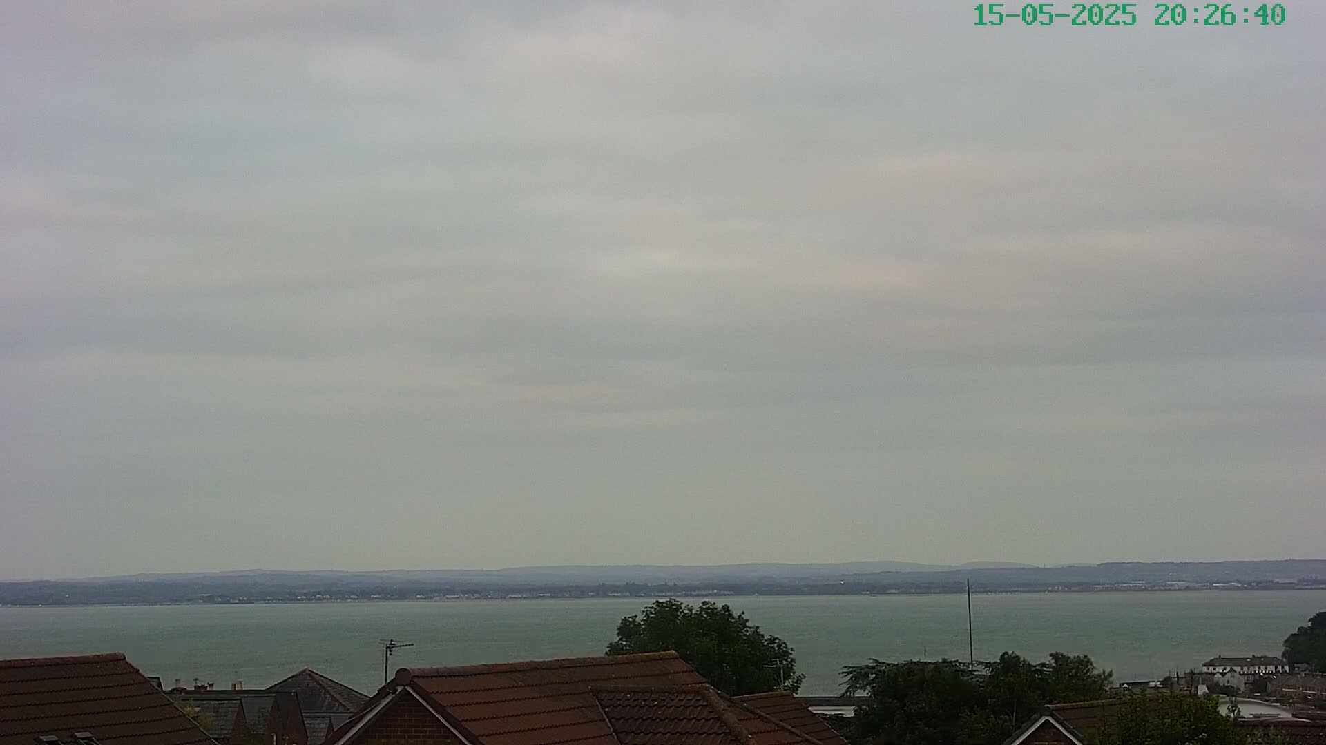 A calm body of water stretches to a distant shoreline under a cloudy sky, viewed from behind the rooftops of houses.