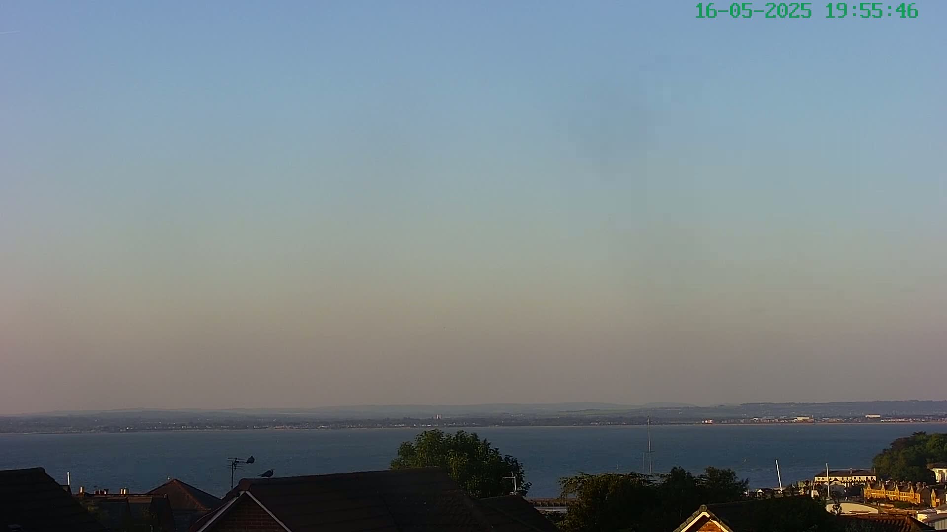 A calm, hazy evening view shows a body of water extending to a distant shoreline, partially obscured by rooftops and trees in the foreground.