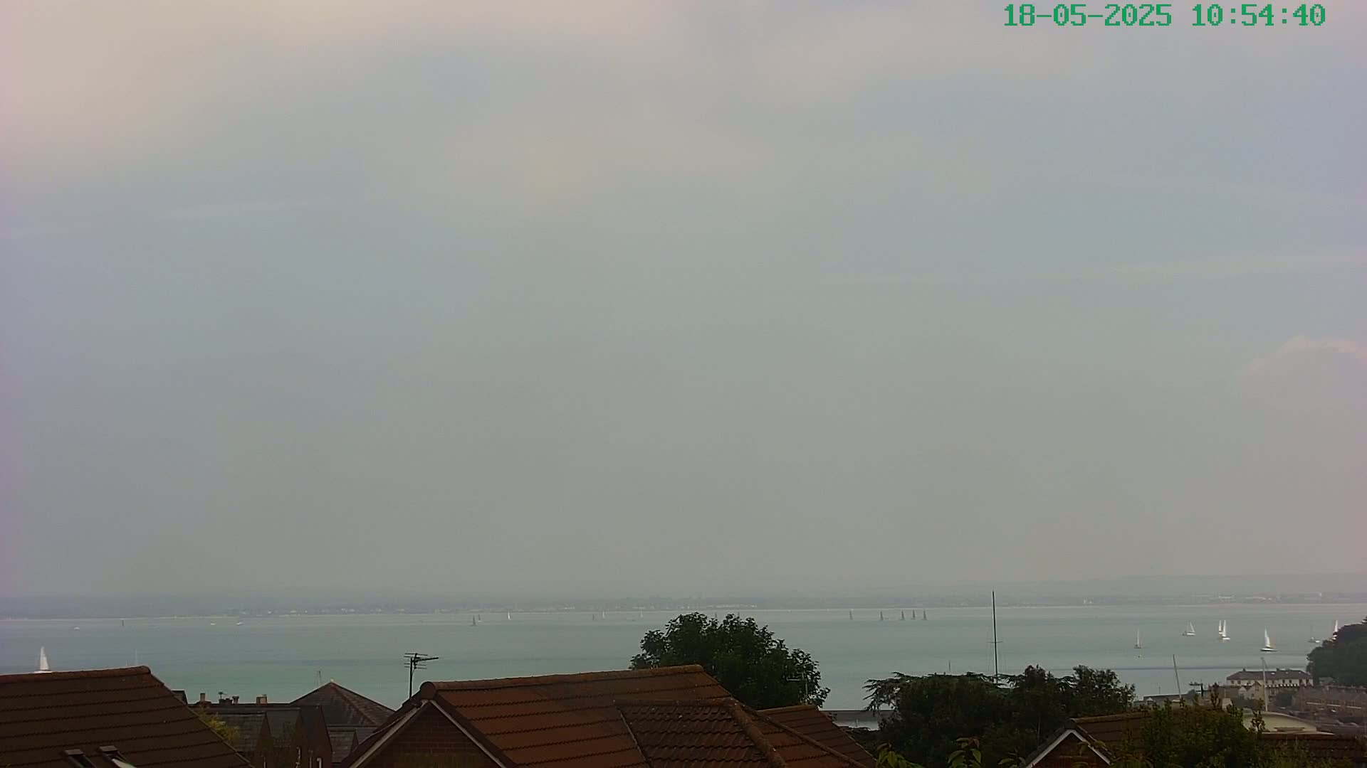 A hazy view of a calm body of water with several sailboats in the distance, seen from behind the brown tiled roofs of houses.