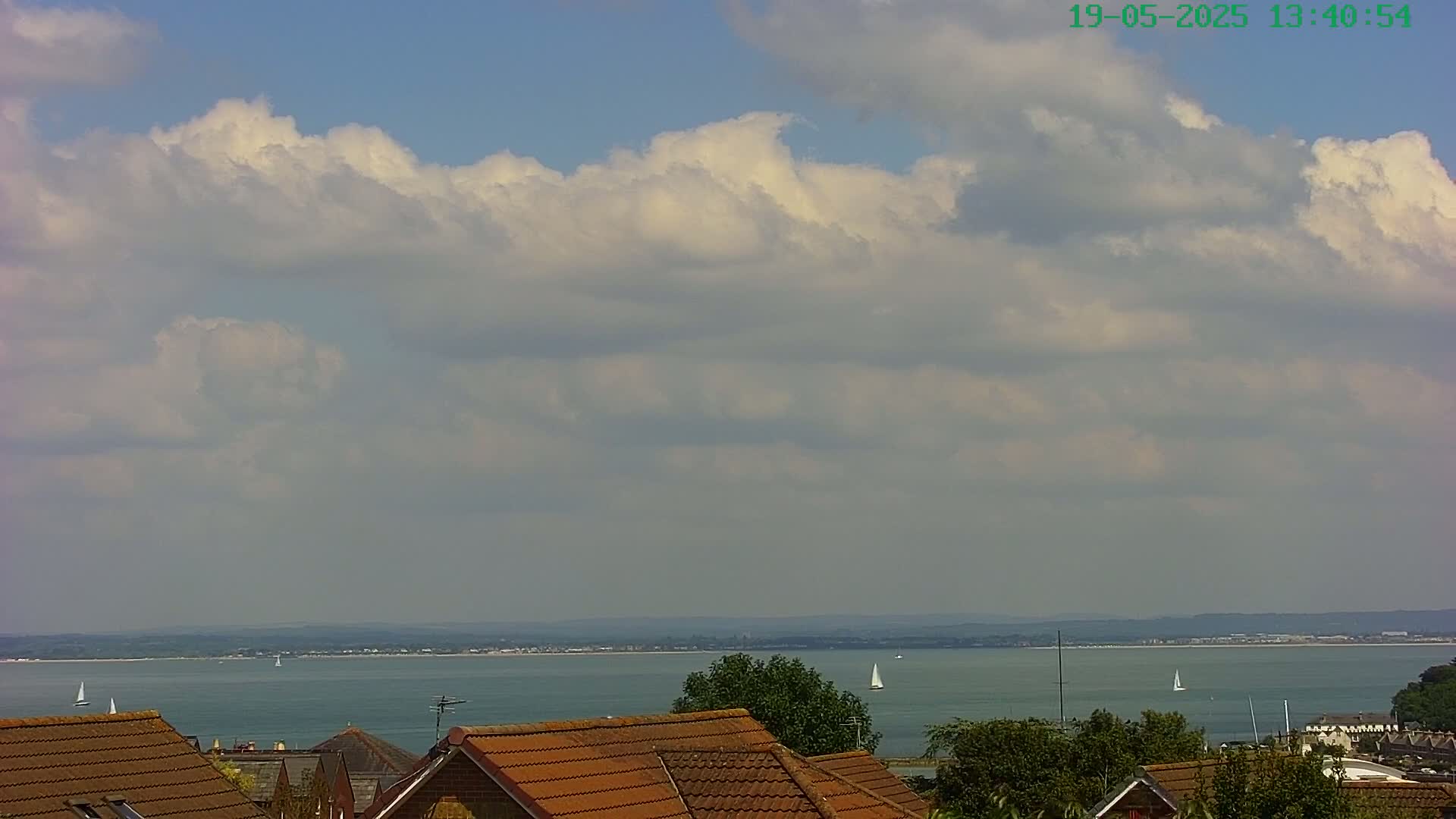 A partly cloudy sky overlooks a calm body of water with a few sailboats visible in the distance, and brown-tiled rooftops are in the foreground.
