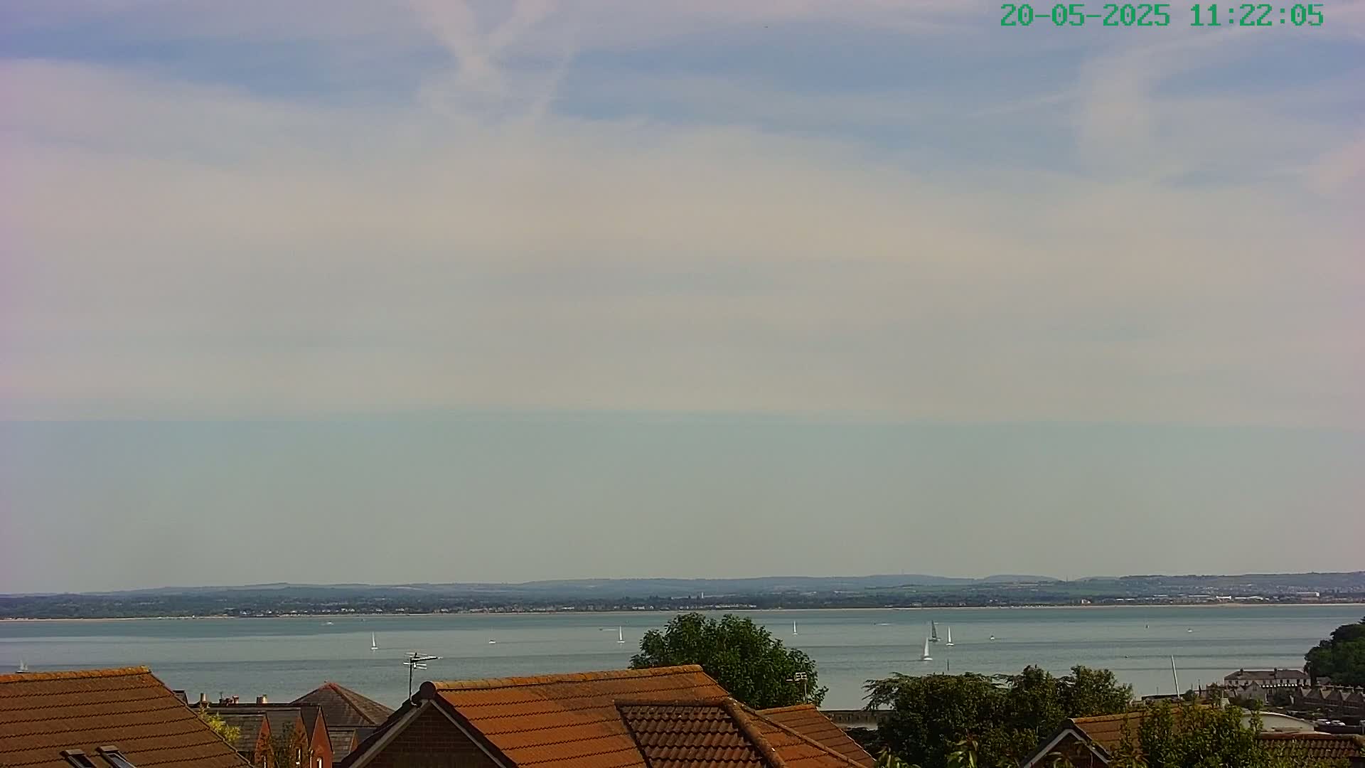 A calm, partly cloudy day overlooks a body of water with several sailboats and distant land, seen from behind a row of brown-tiled rooftops.