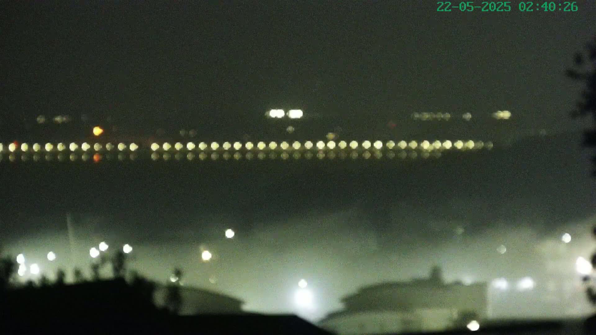 A foggy, nighttime scene shows a line of lights across a body of water, with more lights visible in the foreground and background.