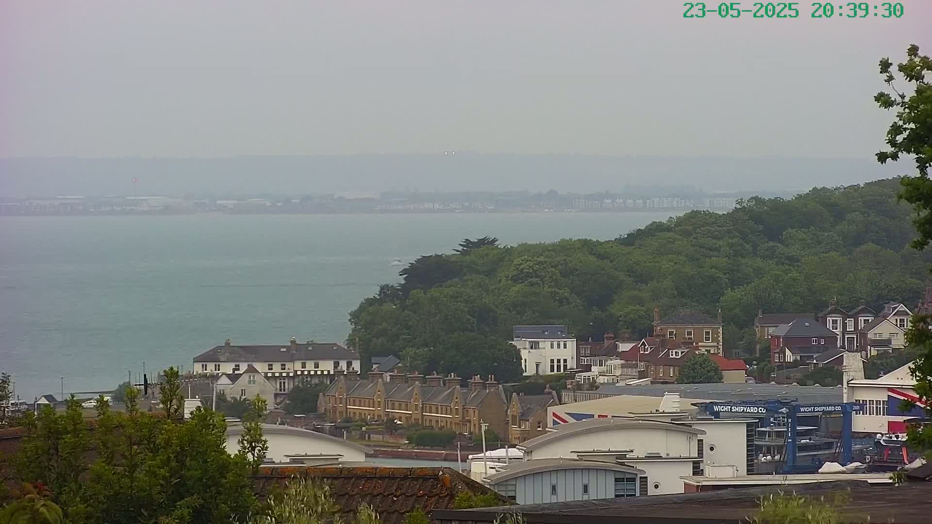 A hazy view of a coastline shows a calm body of water, a green hillside dotted with houses, and a shipyard.