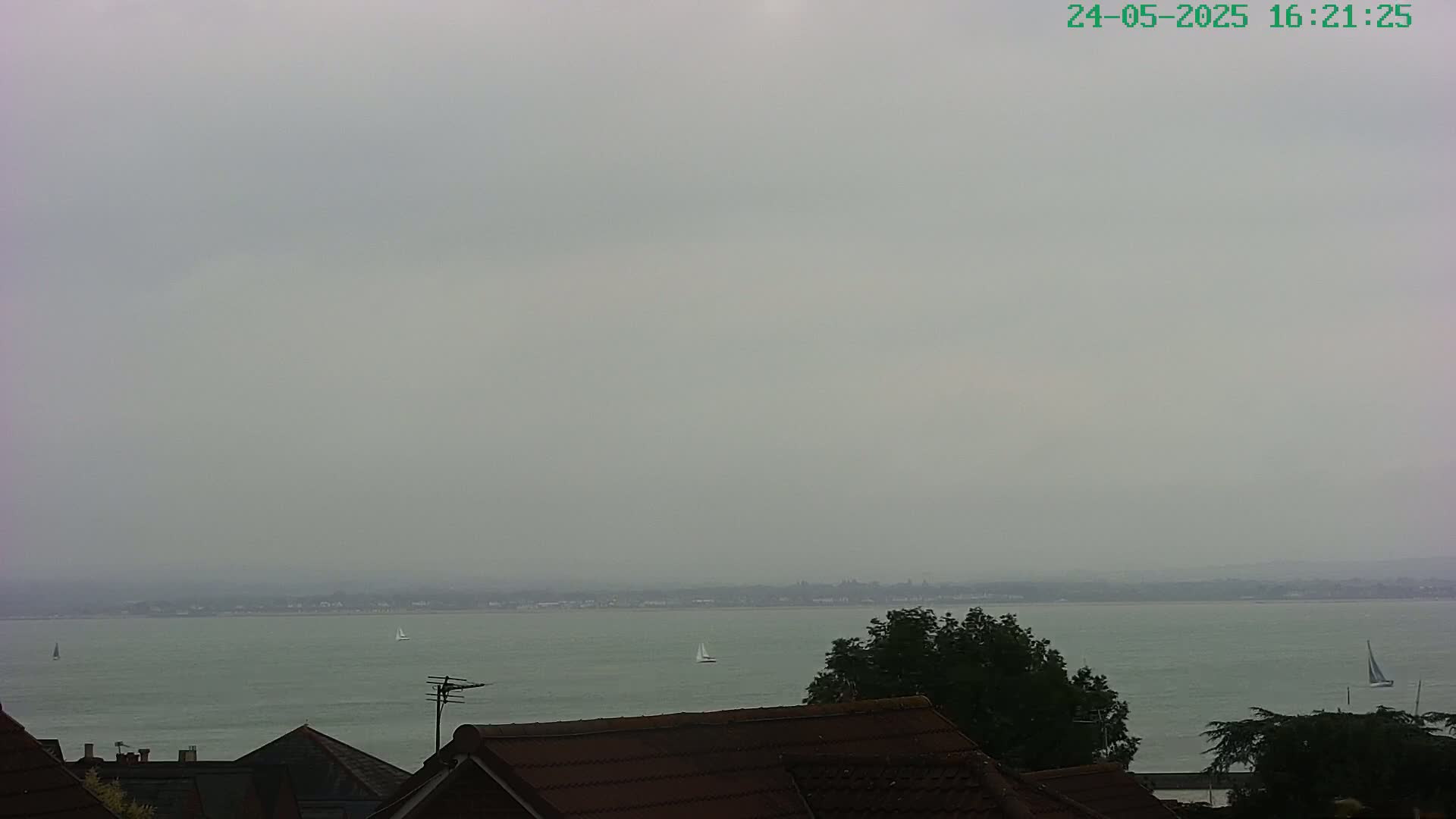 A hazy, overcast day reveals a calm body of water with several sailboats visible in the distance, viewed from atop a row of houses.