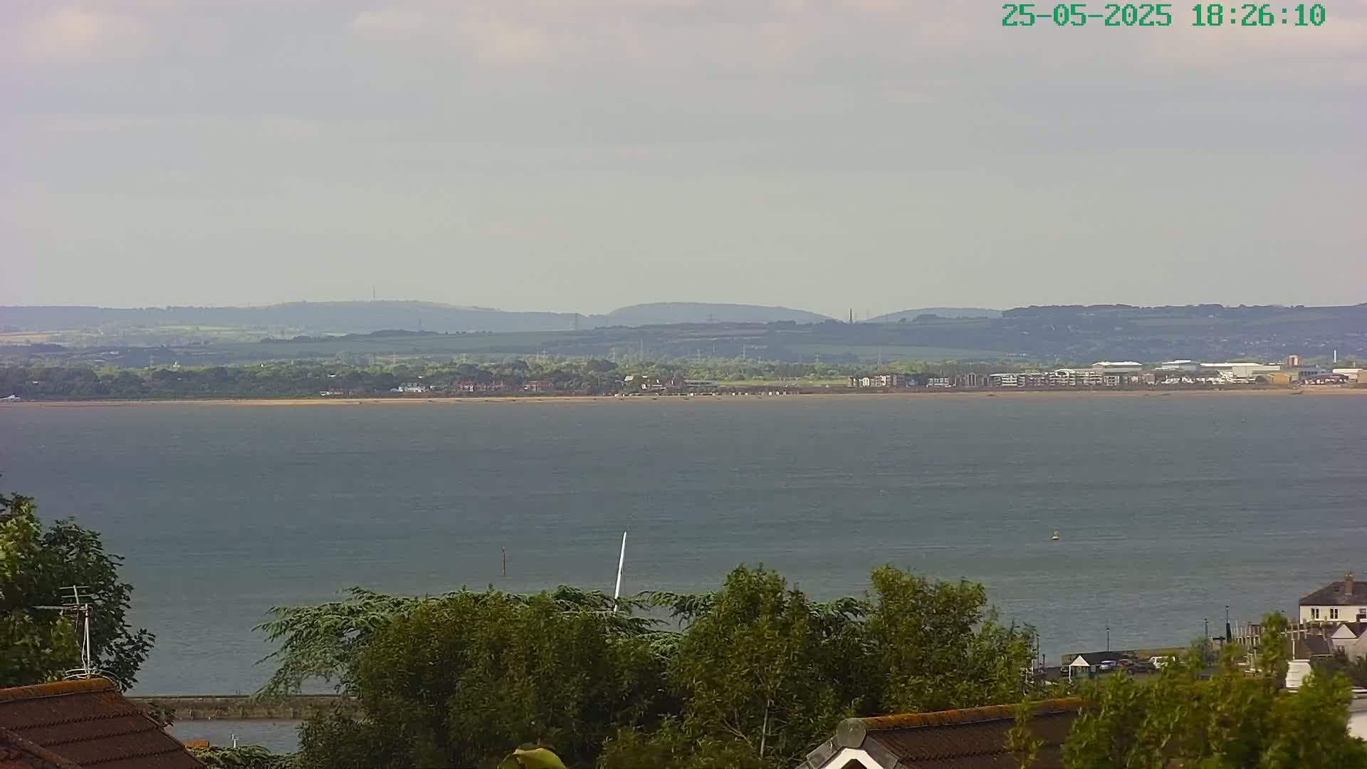A wide shot shows a calm body of water with a distant shoreline featuring buildings and hills under a slightly cloudy sky.