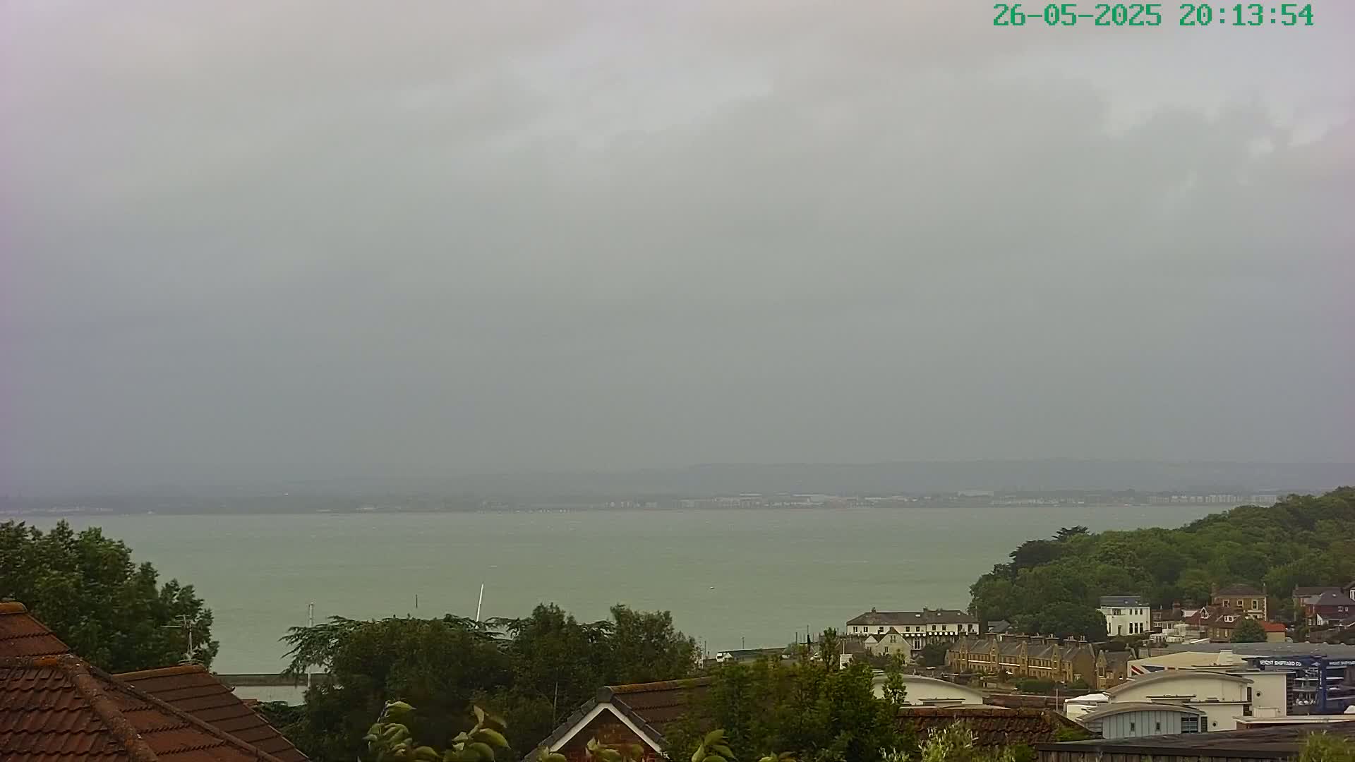 A calm, overcast day reveals a view of a body of water stretching to a distant shoreline, with houses and trees nestled along the foreground.