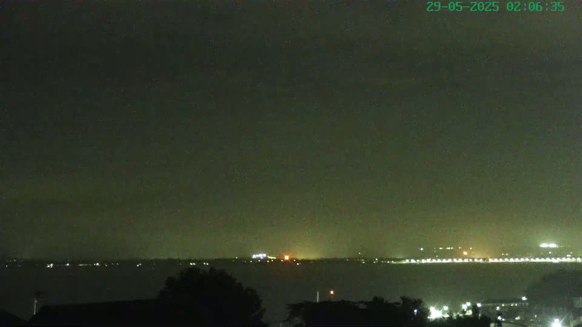 A nighttime view of a body of water with a distant shoreline illuminated by lights under a dark, possibly hazy sky.