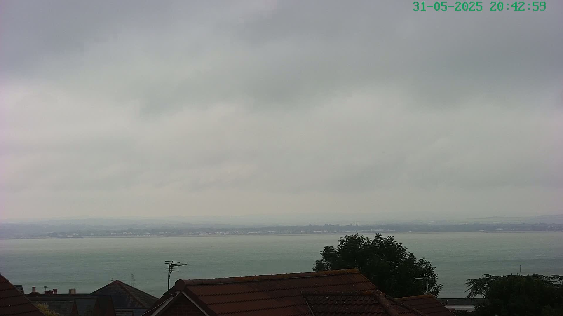 A calm body of water stretches to a hazy horizon under an overcast sky, viewed from behind the rooftops of several houses.