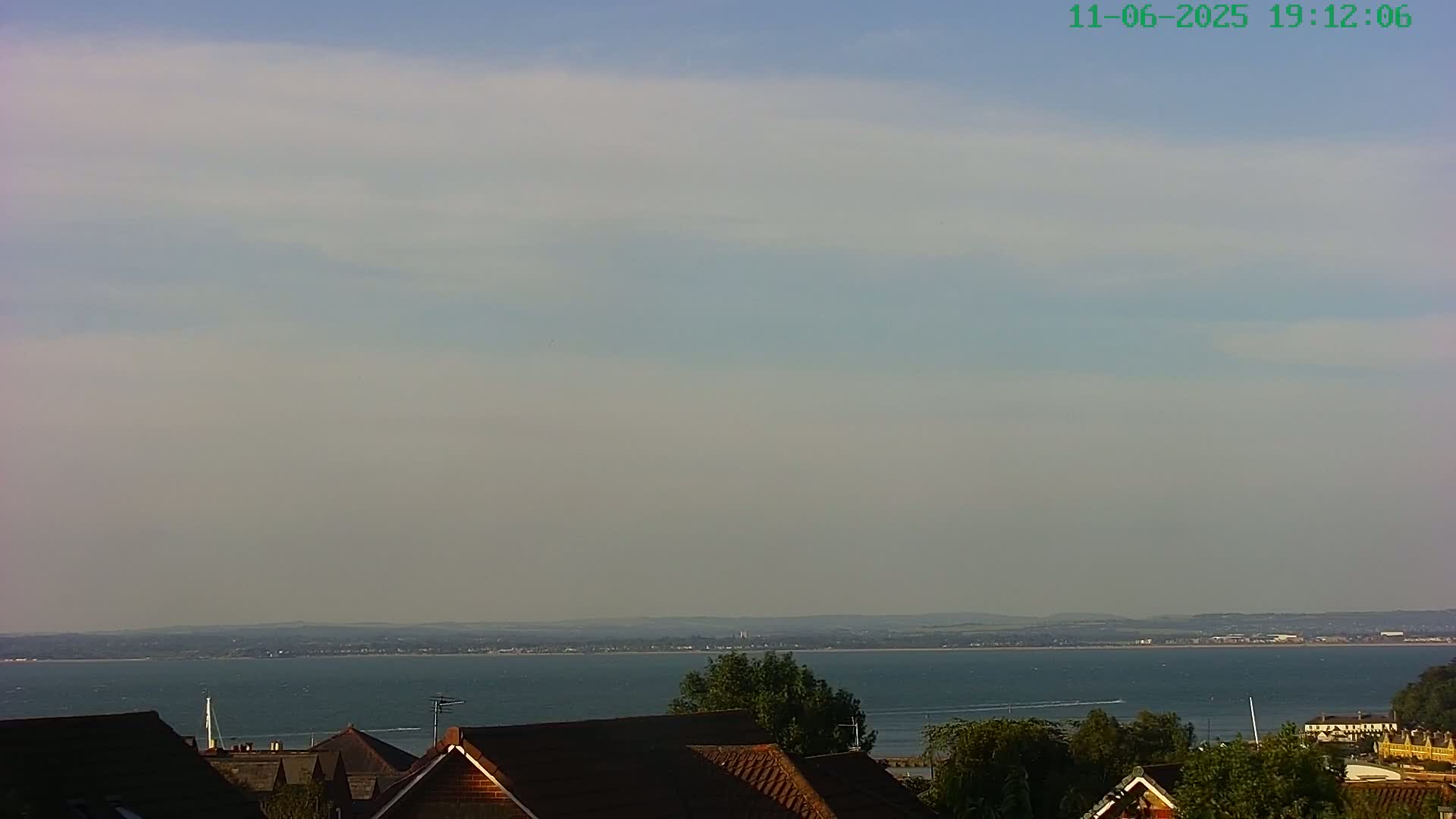 A mostly clear, slightly hazy sky overlooks a calm body of water with a distant shoreline, all viewed from above the rooftops of houses with green trees.