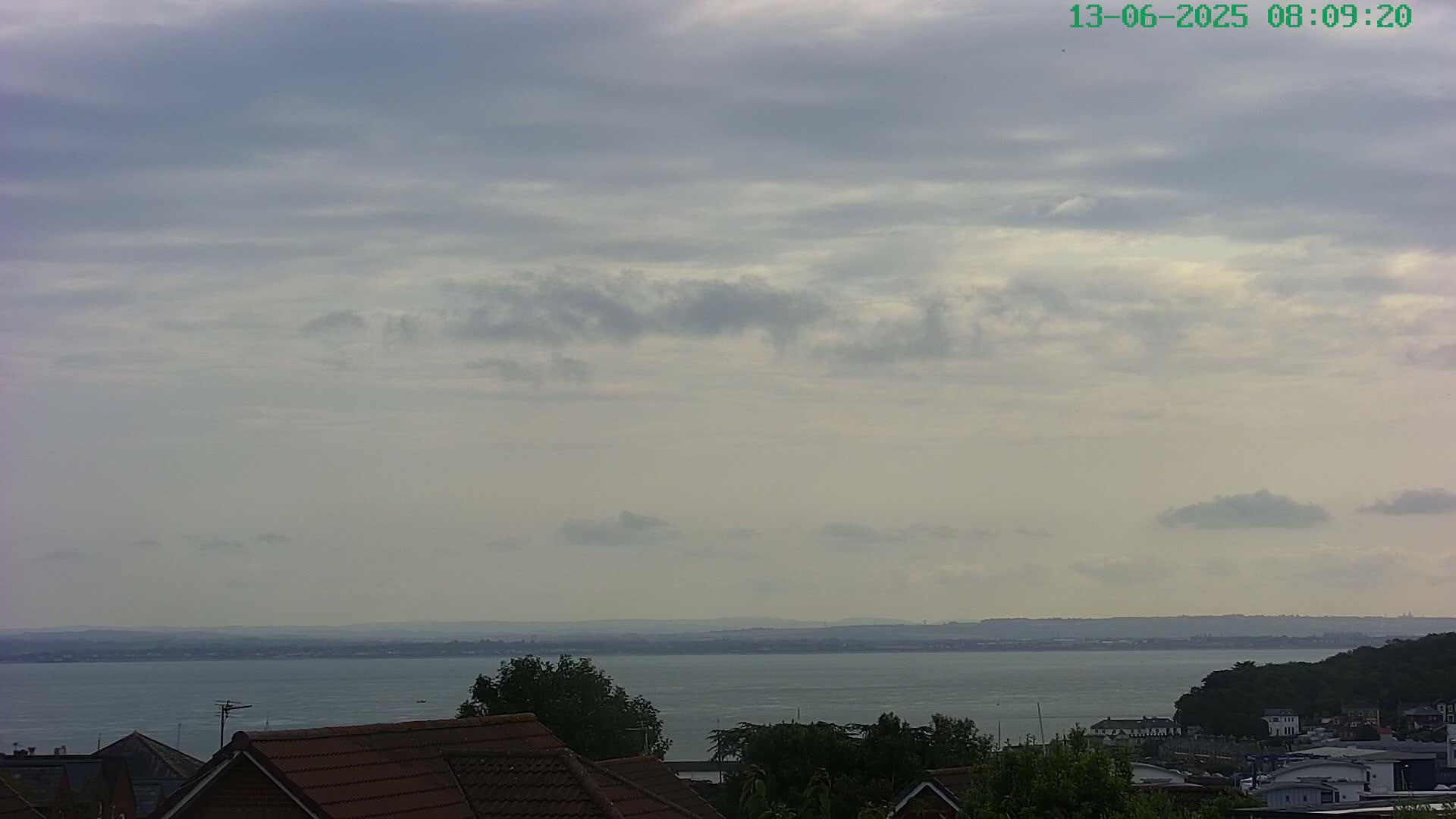 A mostly cloudy sky overlooks a calm body of water and a distant shoreline, with rooftops and green vegetation in the foreground.