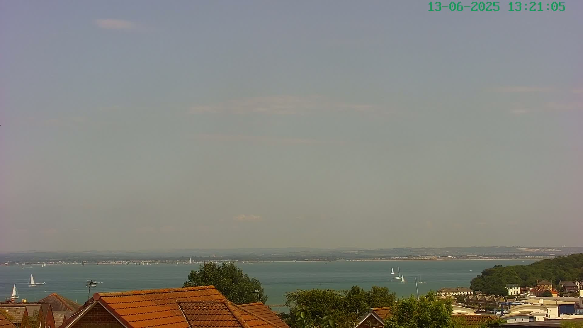 A hazy, sunny day overlooks a calm body of water dotted with sailboats, with a distant shoreline and rooftops of houses in the foreground.