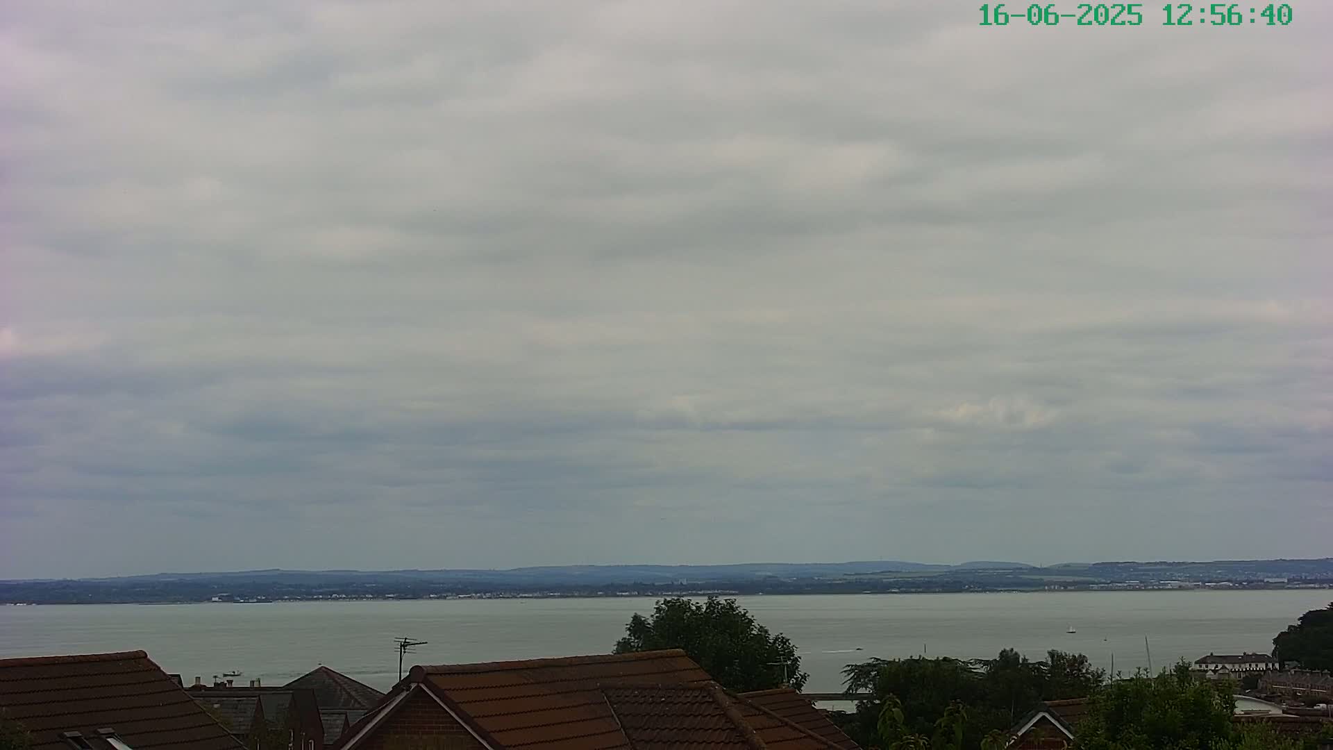 A view of a calm body of water under an overcast sky, seen from behind the rooftops of houses.
