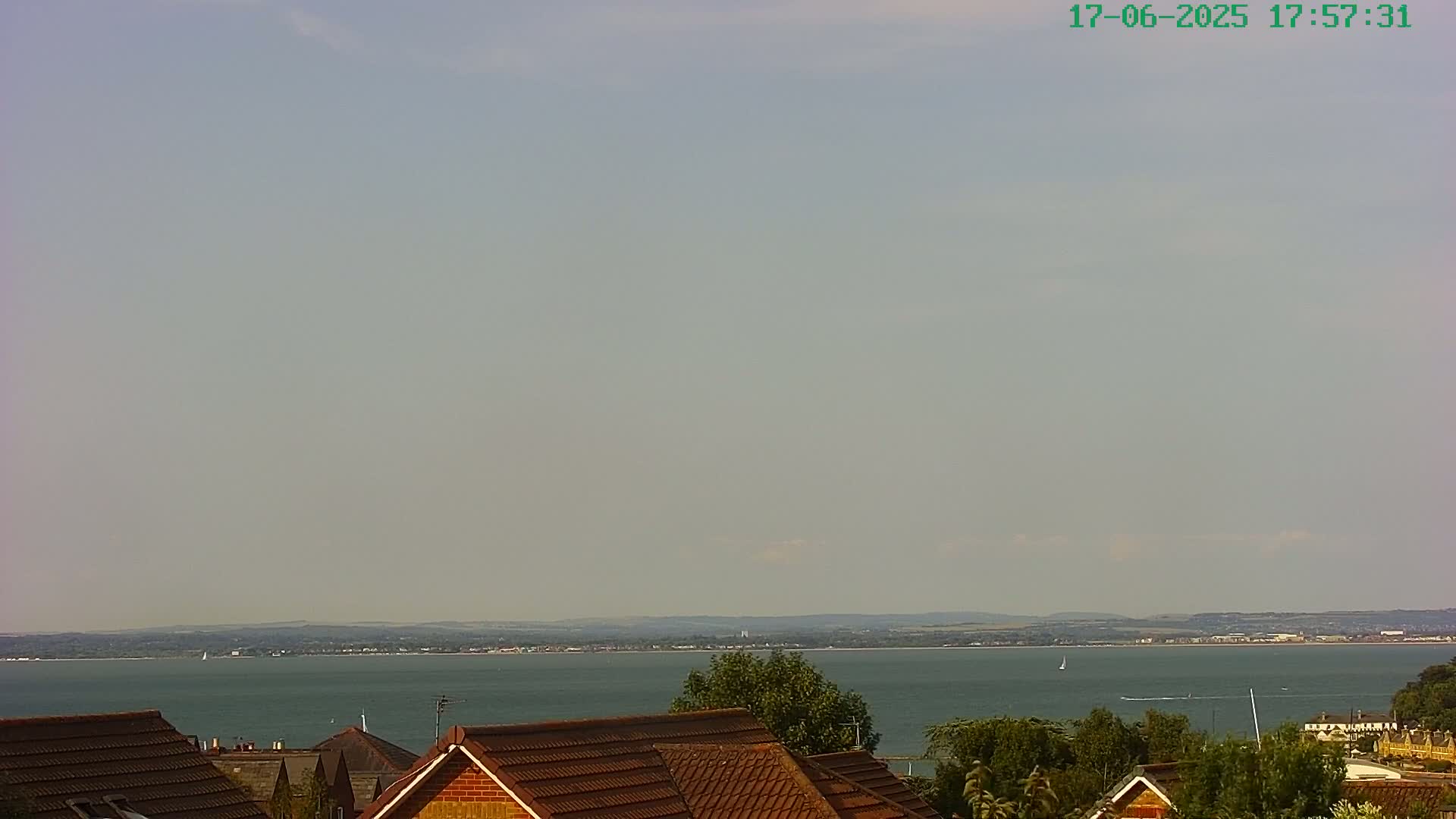 A hazy, sunny day reveals a calm body of water stretching to a distant shoreline, partially obscured by the rooftops of houses in the foreground.